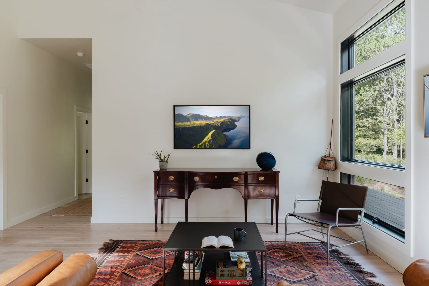 Modern living room with light hardwood floors, a dark wood console table with a TV displaying a landscape, and a black metal and leather armchair, all illuminated by large windows overlooking a forest.
