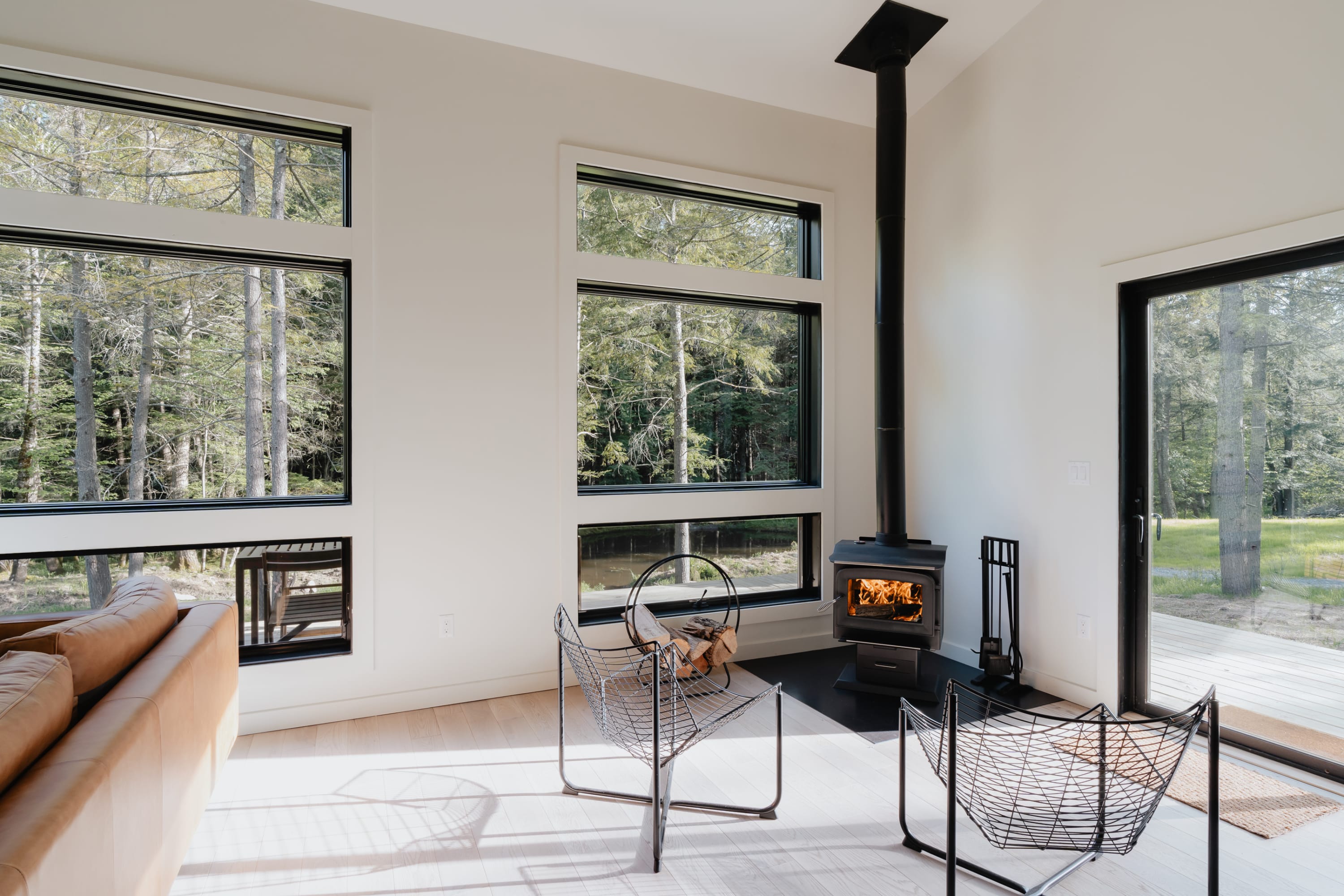 Bright living area featuring a black wood-burning stove with stacked firewood, two modern wire chairs, and a brown leather sofa. Multiple large windows provide abundant natural light and views of the surrounding forest.