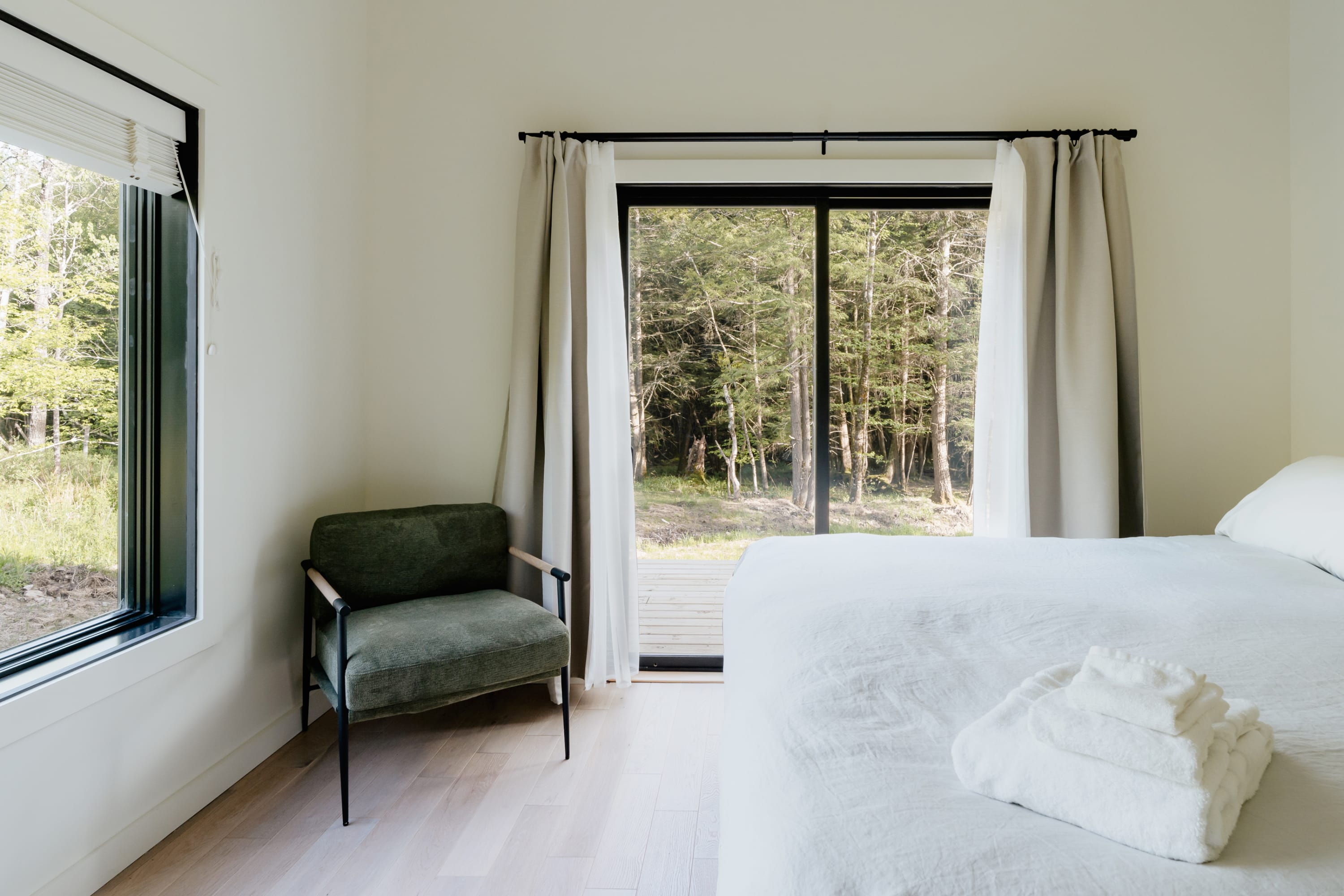 Serene bedroom with a white bed, light hardwood floors, and a green accent chair, featuring a large sliding glass door leading to an outdoor area and a window offering forest views.