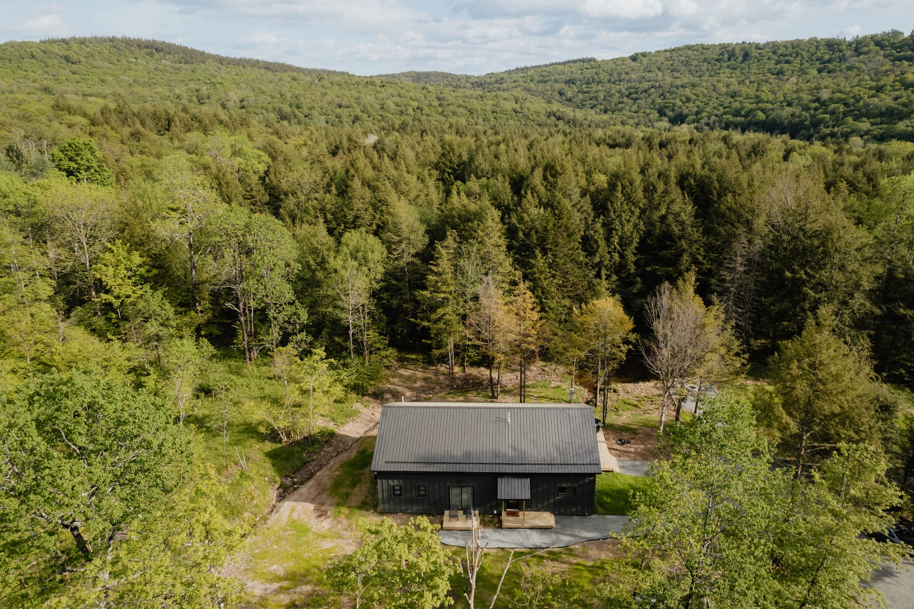 Aerial view of a dark gray modern cabin nestled in a vast, lush green forest with rolling hills in the background. The cabin has a metal roof and is surrounded by trees, highlighting its secluded natural setting.
