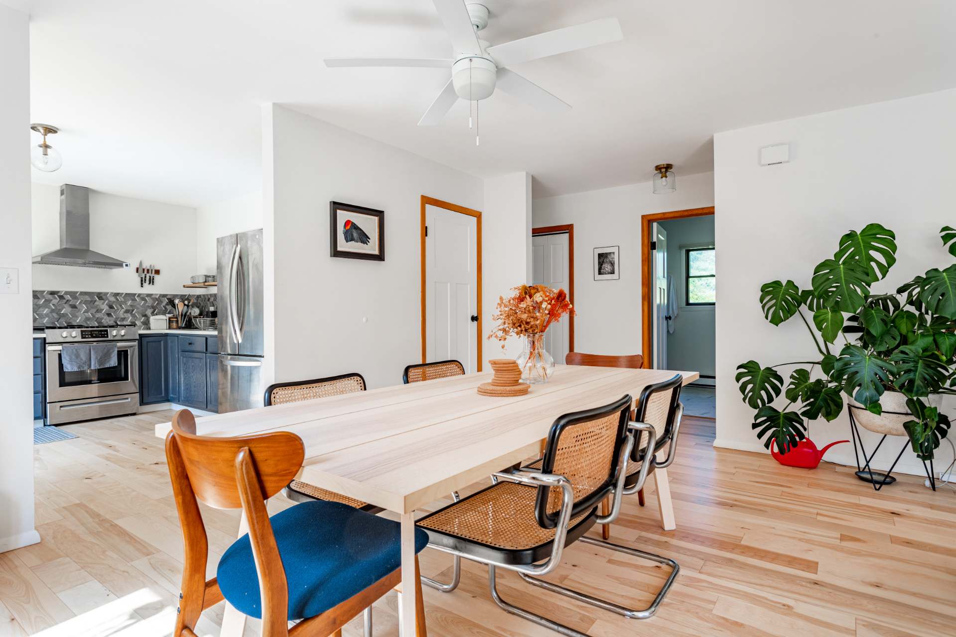 Spacious dining area with a light wood table and a mix of modern and cane-back chairs, set on light hardwood floors. A large Monstera plant adds a touch of nature, and a white ceiling fan hangs overhead.