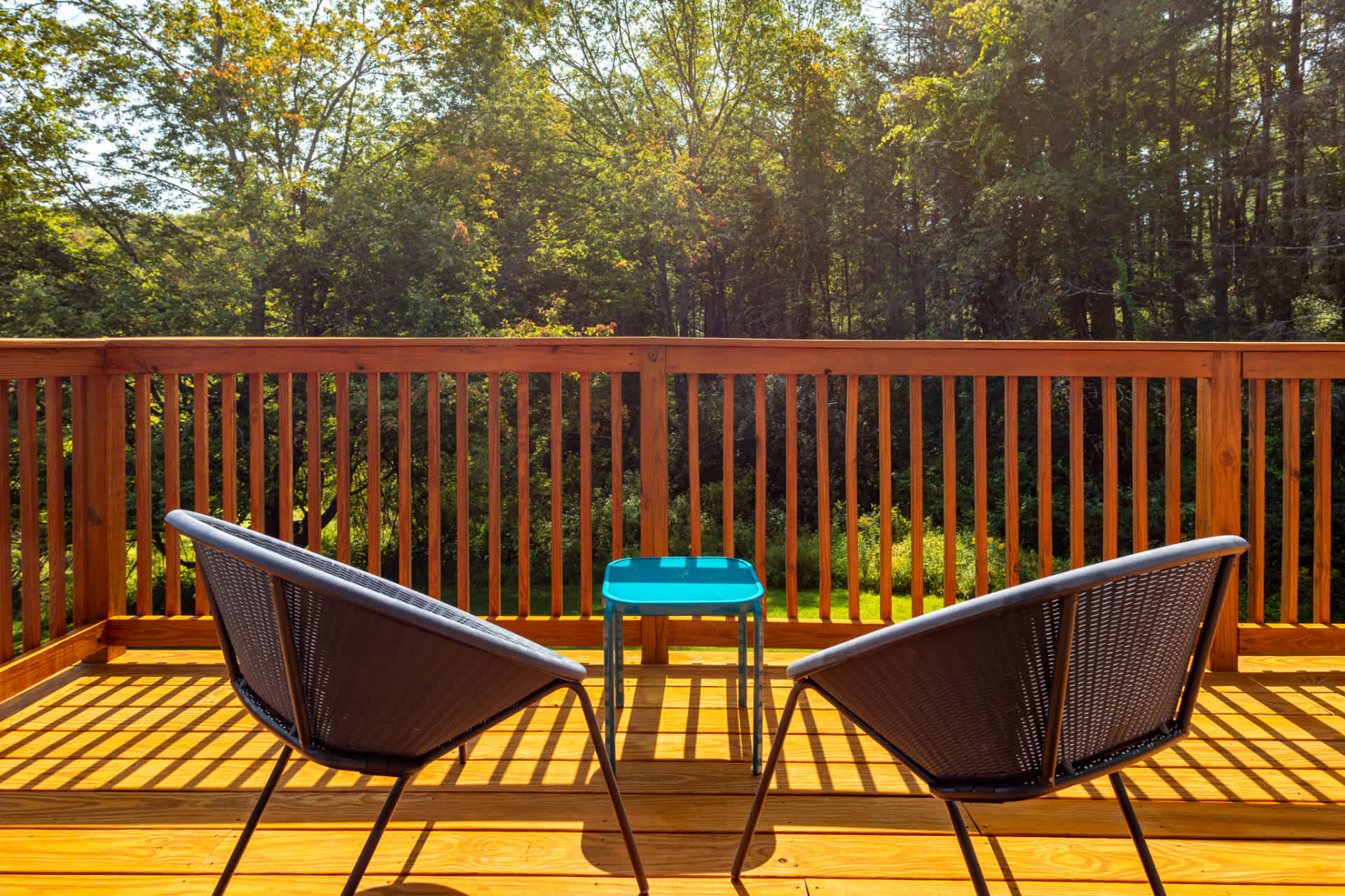 Close-up view of a wooden balcony with two modern dark grey chairs and a small blue side table, offering a serene view of lush green trees and foliage under bright sunlight.