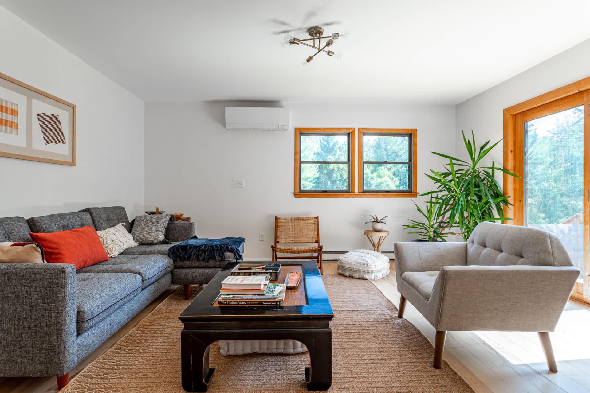 Modern living room with a grey sectional sofa adorned with colorful pillows, a dark wooden coffee table, a light grey armchair, and a large potted plant, all on a natural fiber rug with light wood floors.