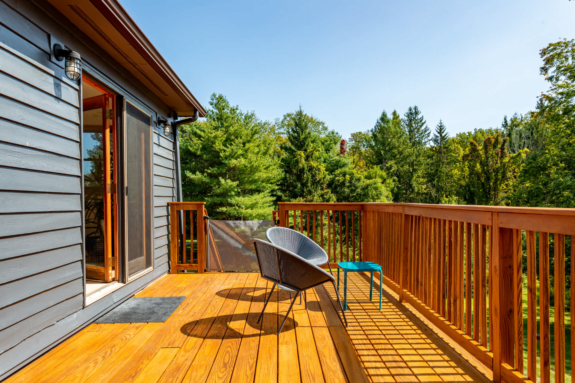 A spacious wooden deck with two modern outdoor chairs and a small blue side table, offering a view of lush green trees under a clear blue sky. A sliding glass door leads into the house.