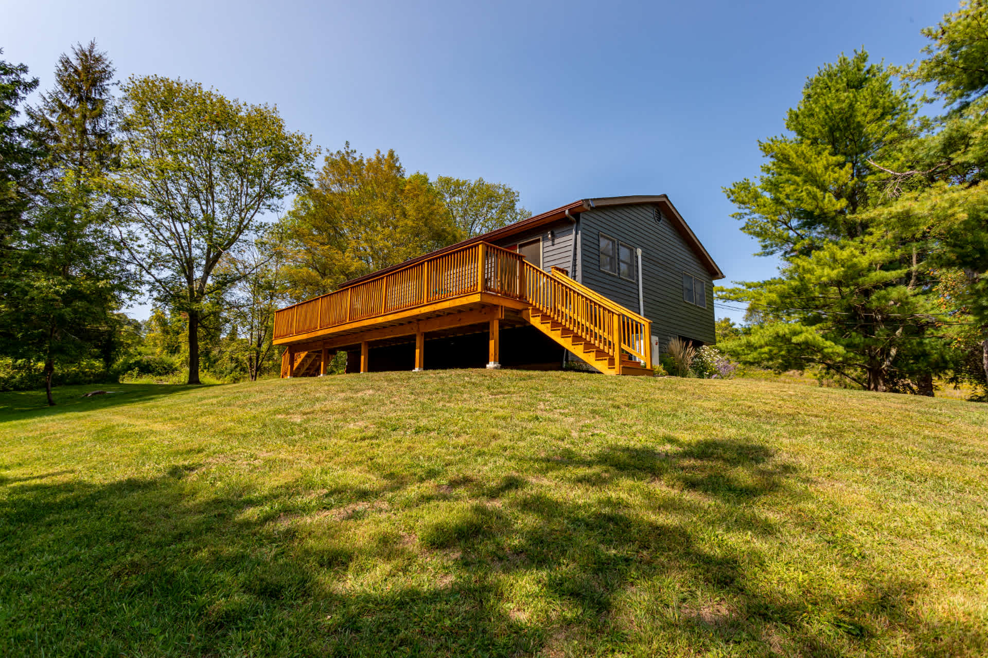 Ground-level view of the dark gray house with a prominent wooden deck and stairs, situated on a gentle grassy slope surrounded by mature green trees under a clear blue sky. The deck offers ample space for outdoor seating.