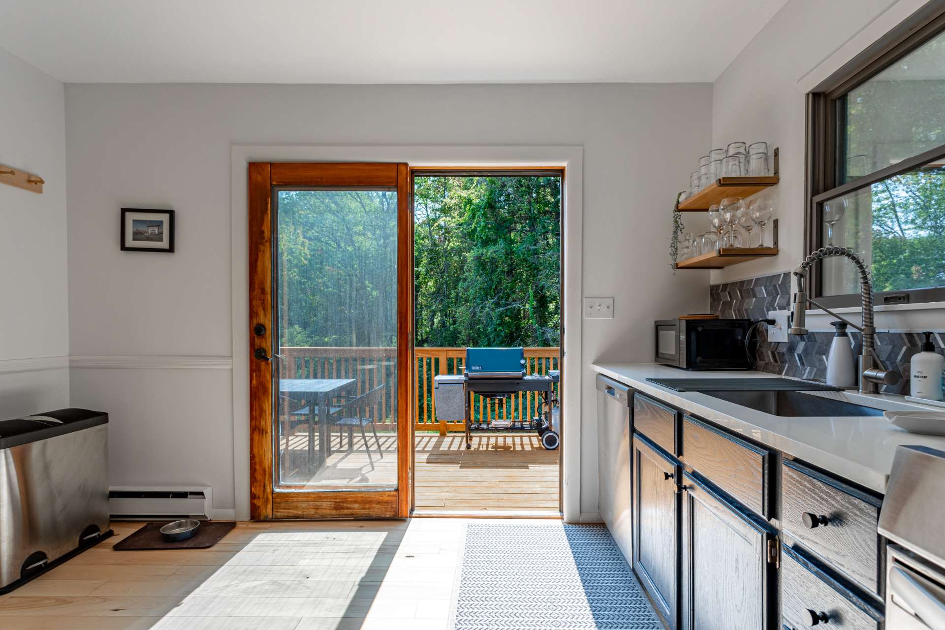 Bright kitchen area with a stainless steel sink, white countertops, and dark blue cabinets, featuring a sliding glass door that opens to an outdoor deck with a BBQ grill. Open wooden shelves hold glassware, and a microwave is visible.