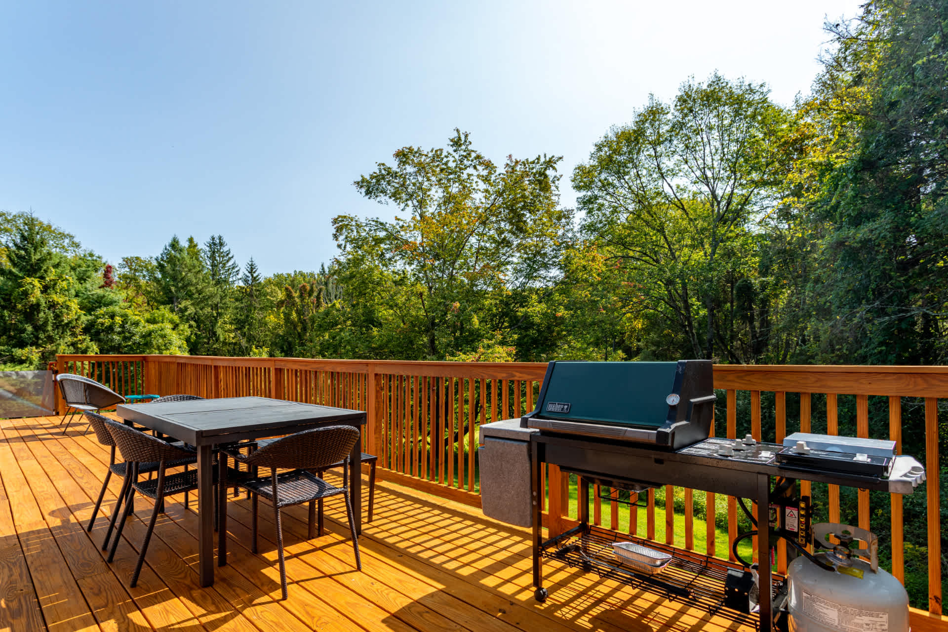 Spacious wooden deck with a dark dining table and chairs, alongside a large black BBQ grill, all set against a backdrop of dense green trees under a clear blue sky.