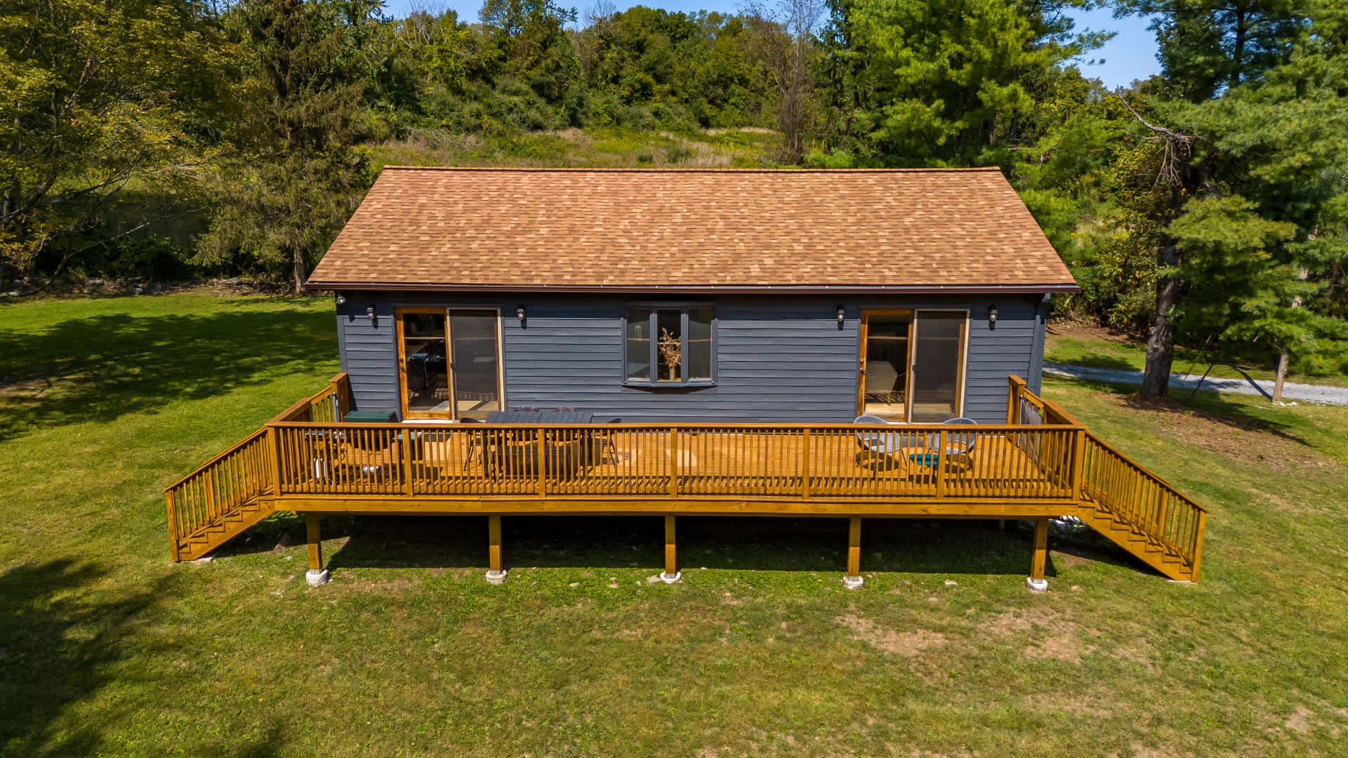 Aerial view of a dark gray house with a brown roof, featuring a large wooden deck with railings and two sets of stairs leading down to a grassy lawn. The property is surrounded by lush green trees under a bright sky.