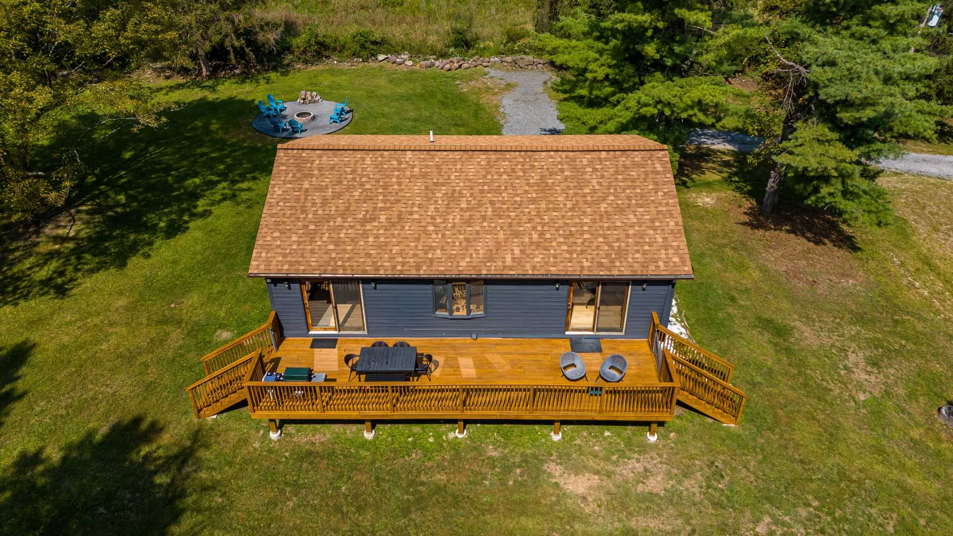 Aerial view of a dark blue house with a brown shingle roof, featuring a large wooden deck with outdoor seating. A fire pit area with blue Adirondack chairs is visible in the grassy yard, surrounded by mature trees.