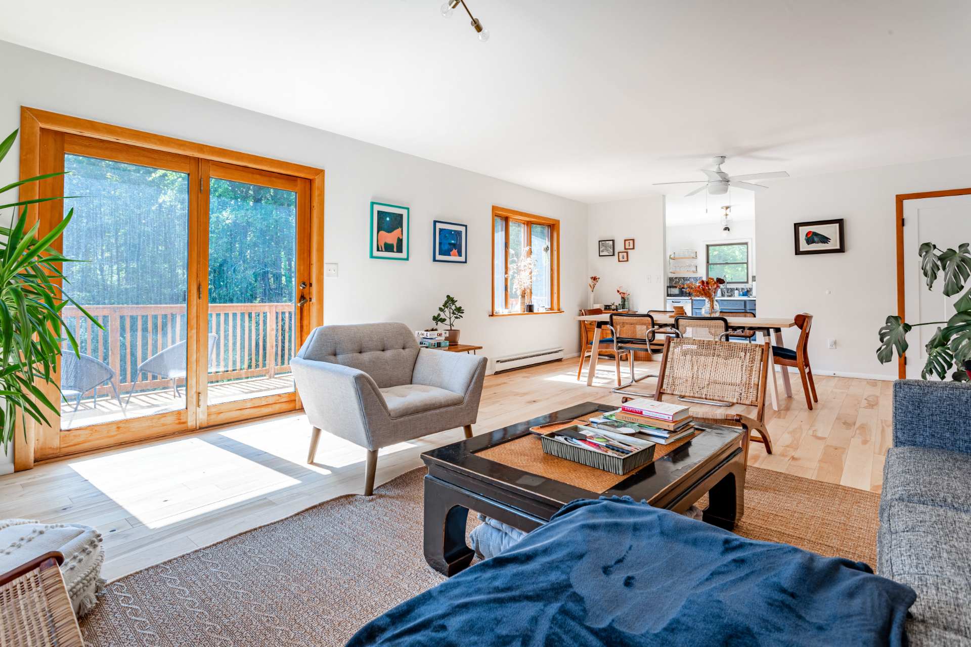 Sunlit living room with light hardwood floors and white walls, featuring a light gray armchair and large wooden sliding glass doors leading to an outdoor deck. Artwork adorns the walls, and a small plant sits on a side table.