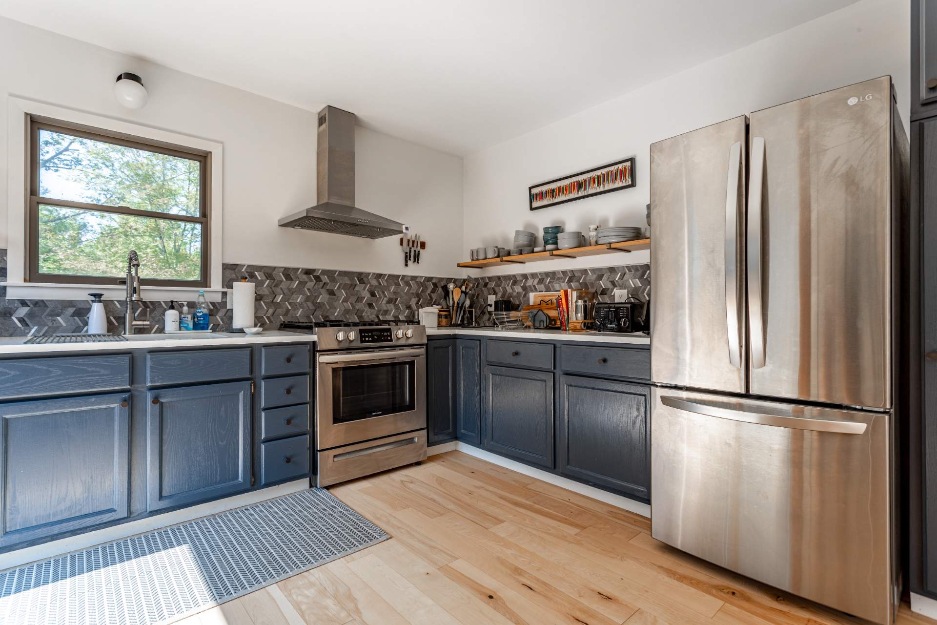 Modern kitchen featuring dark blue cabinets, white countertops, and a striking gray geometric tile backsplash. Stainless steel appliances include a gas range with a hood and a large French door refrigerator, all set on light hardwood floors.