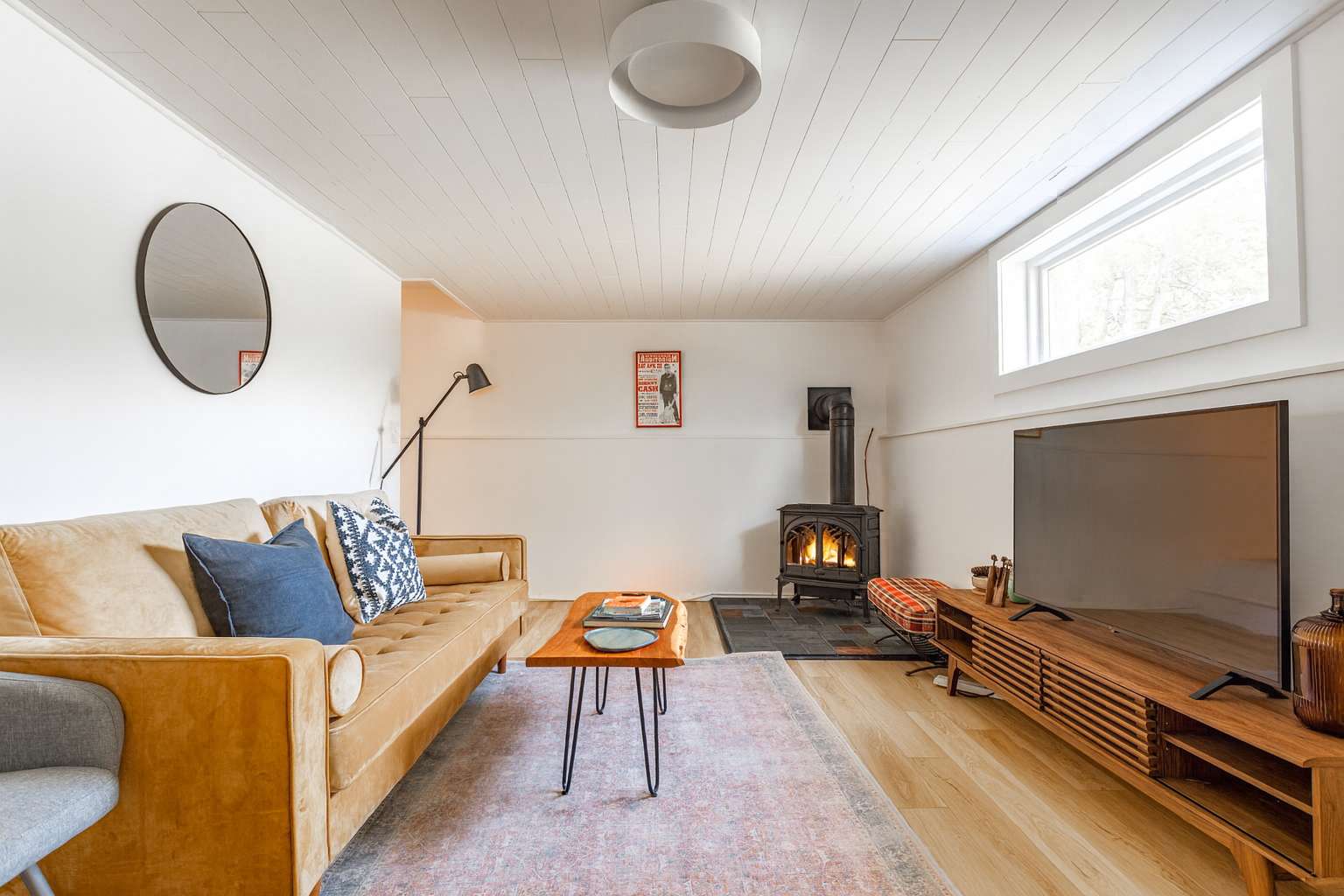 Cozy living room featuring a tan tufted sofa with blue and patterned pillows, a black wood-burning stove on a slate hearth, a wooden coffee table, and a large flat-screen TV on a mid-century modern console, all on light wood floors.