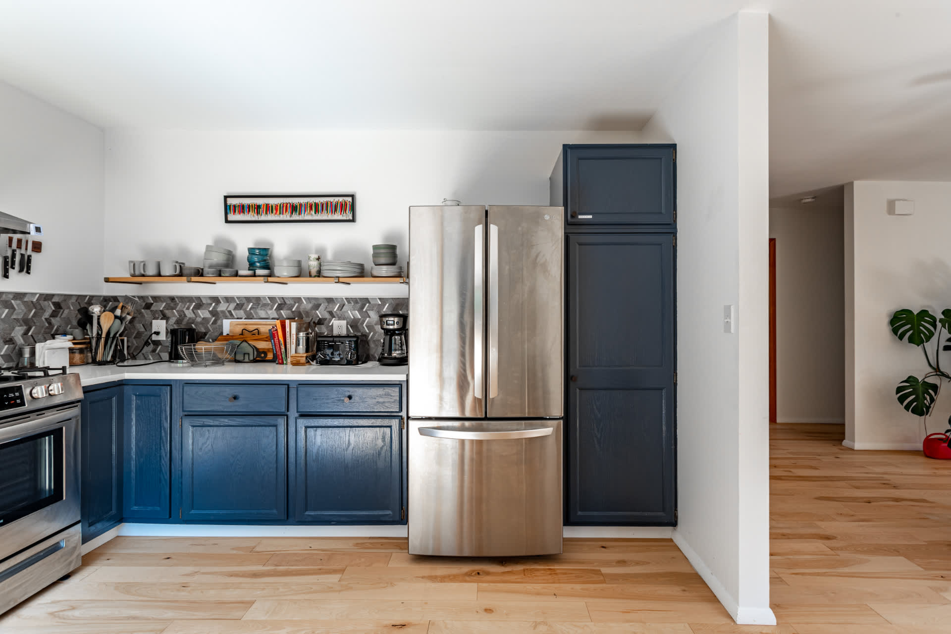 Contemporary kitchen with dark blue base cabinets, white countertops, and a gray geometric tile backsplash, complemented by open wooden shelving. A large stainless steel French door refrigerator stands next to a tall pantry cabinet.