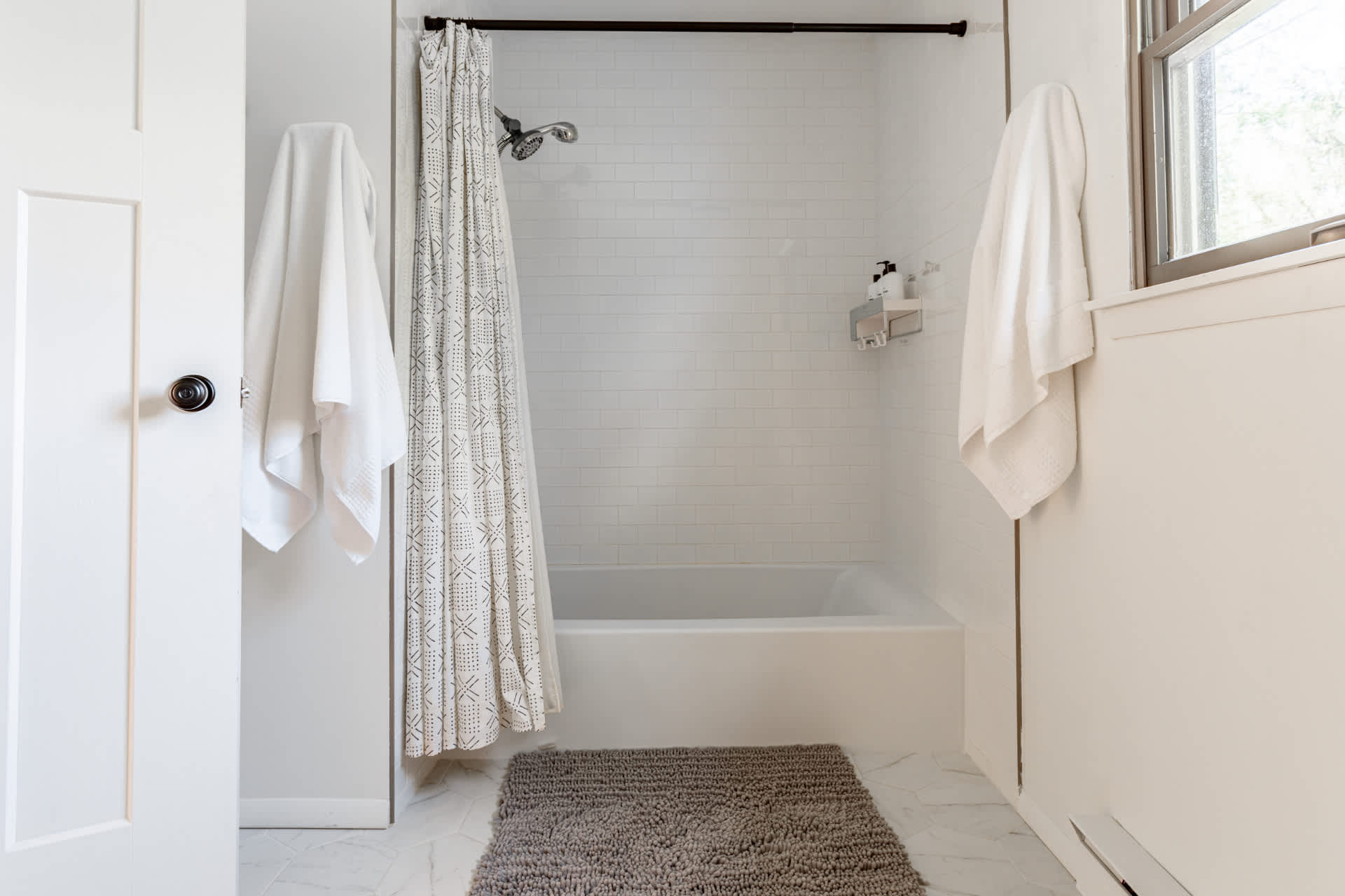 A bright bathroom with a white subway-tiled shower/bathtub combo, a patterned shower curtain, and a dark bath mat on light-colored floor tiles. White towels hang on the wall.