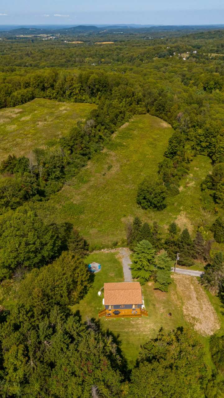 High aerial view showcasing the dark gray house with its brown roof and wooden deck, nestled amidst vast expanses of green forests and fields, with distant mountains under a clear sky. A winding driveway leads to the secluded property.