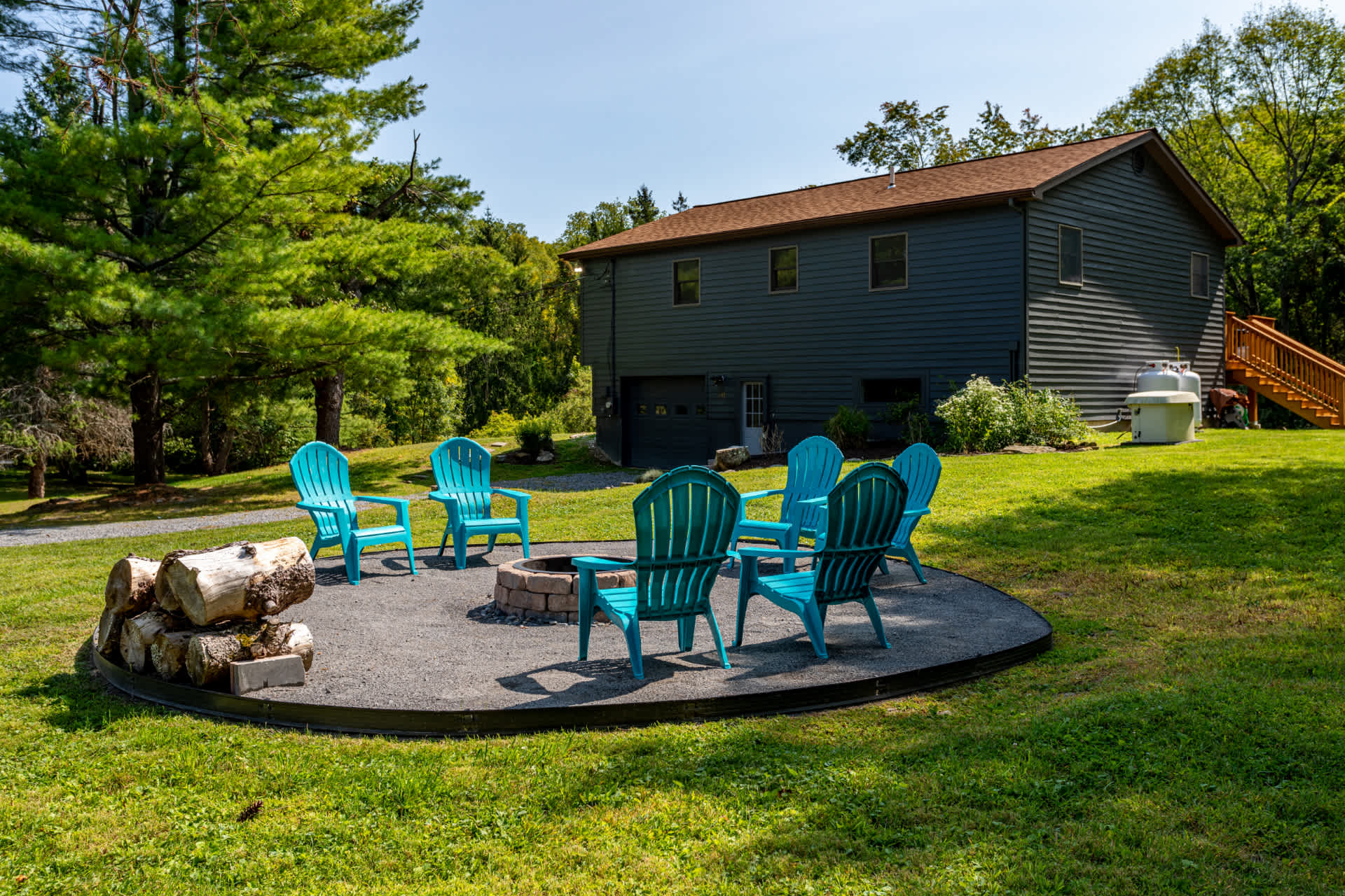 The back exterior of a dark blue house with a brown roof, overlooking a large grassy yard with a circular fire pit area and blue Adirondack chairs. Mature trees surround the property.