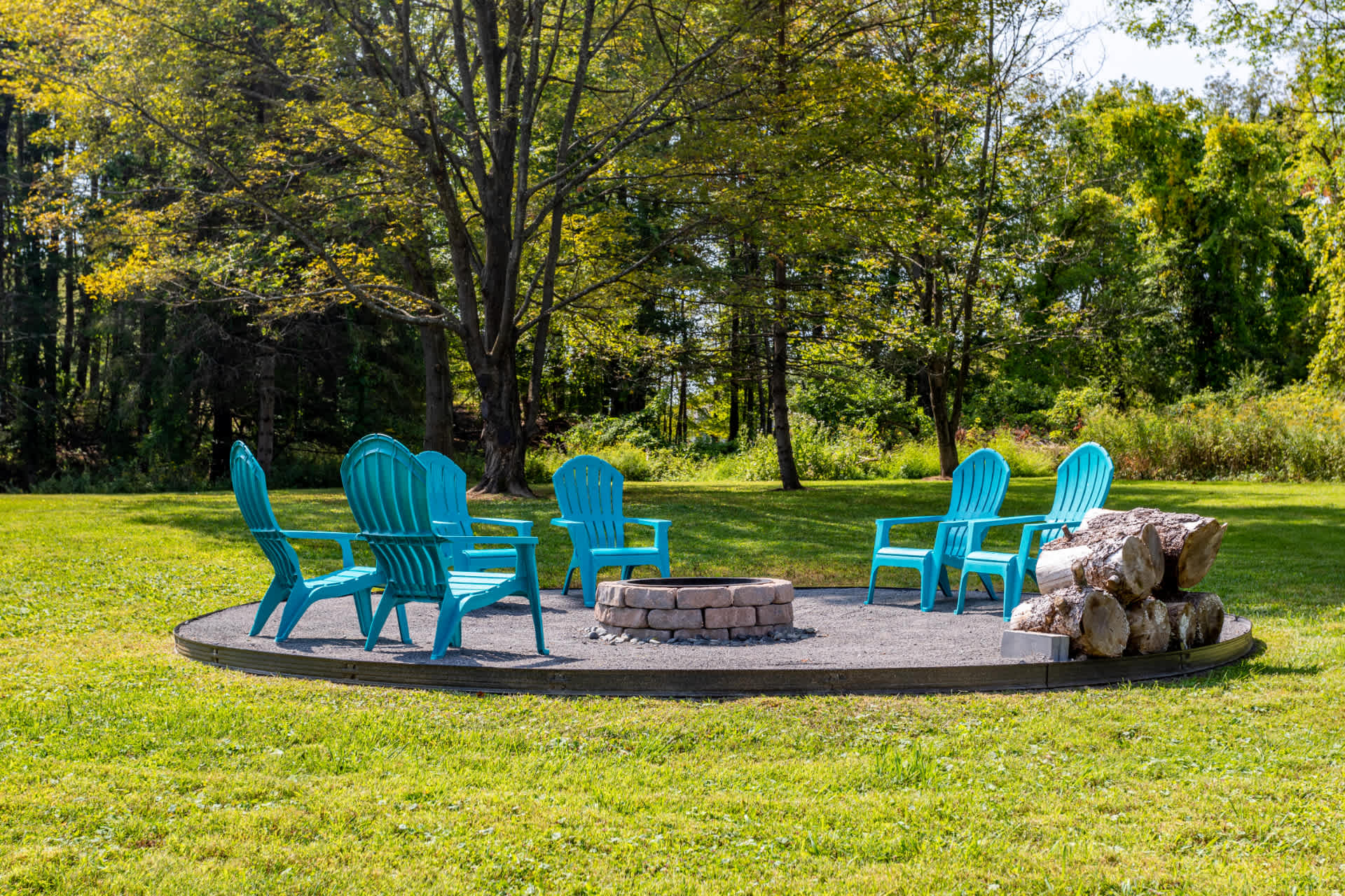 Outdoor fire pit area with a stone ring, surrounded by six bright blue Adirondack chairs, set on a circular gravel base in a grassy yard, with mature trees in the background under a sunny sky.