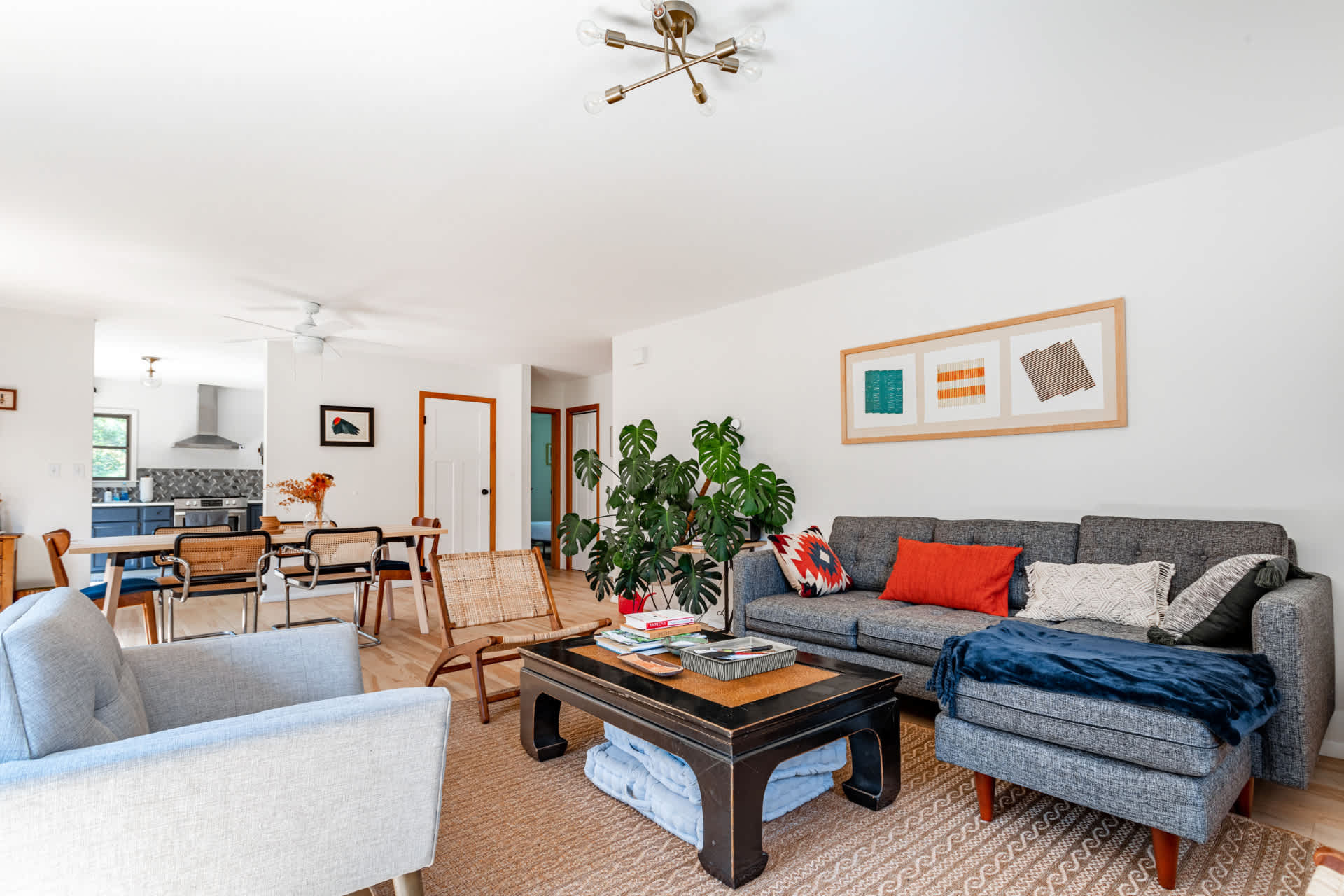 Open-concept living space featuring a gray sectional sofa, a dark coffee table on a textured rug, and light hardwood floors. The room flows into a dining area, with a large Monstera plant and a modern ceiling light fixture.