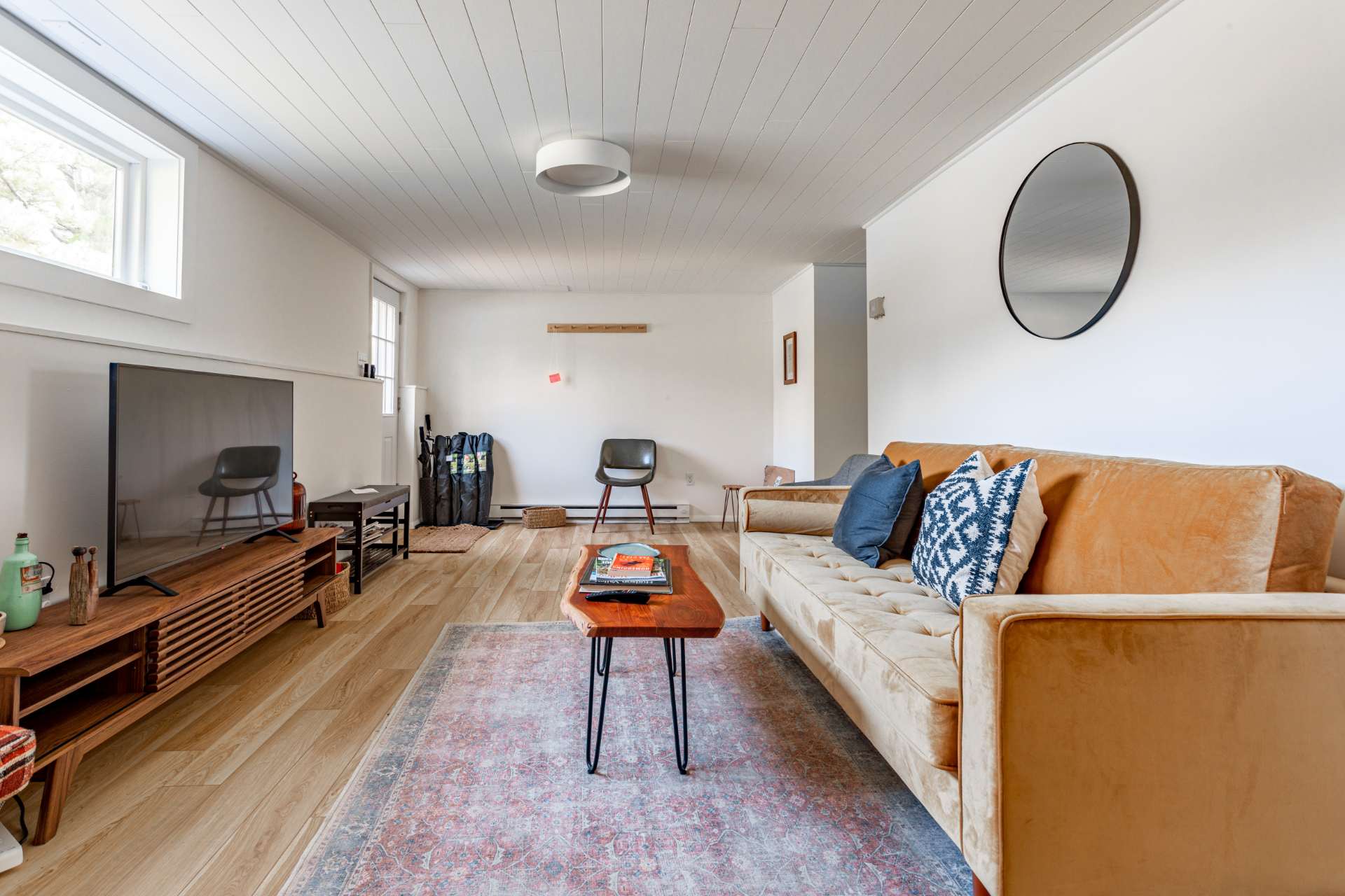 Cozy secondary living space with a long tan velvet sofa and a rustic wood coffee table on a patterned rug, facing a large flat-screen TV on a wooden console. The room features white planked ceilings and light wood-look flooring.