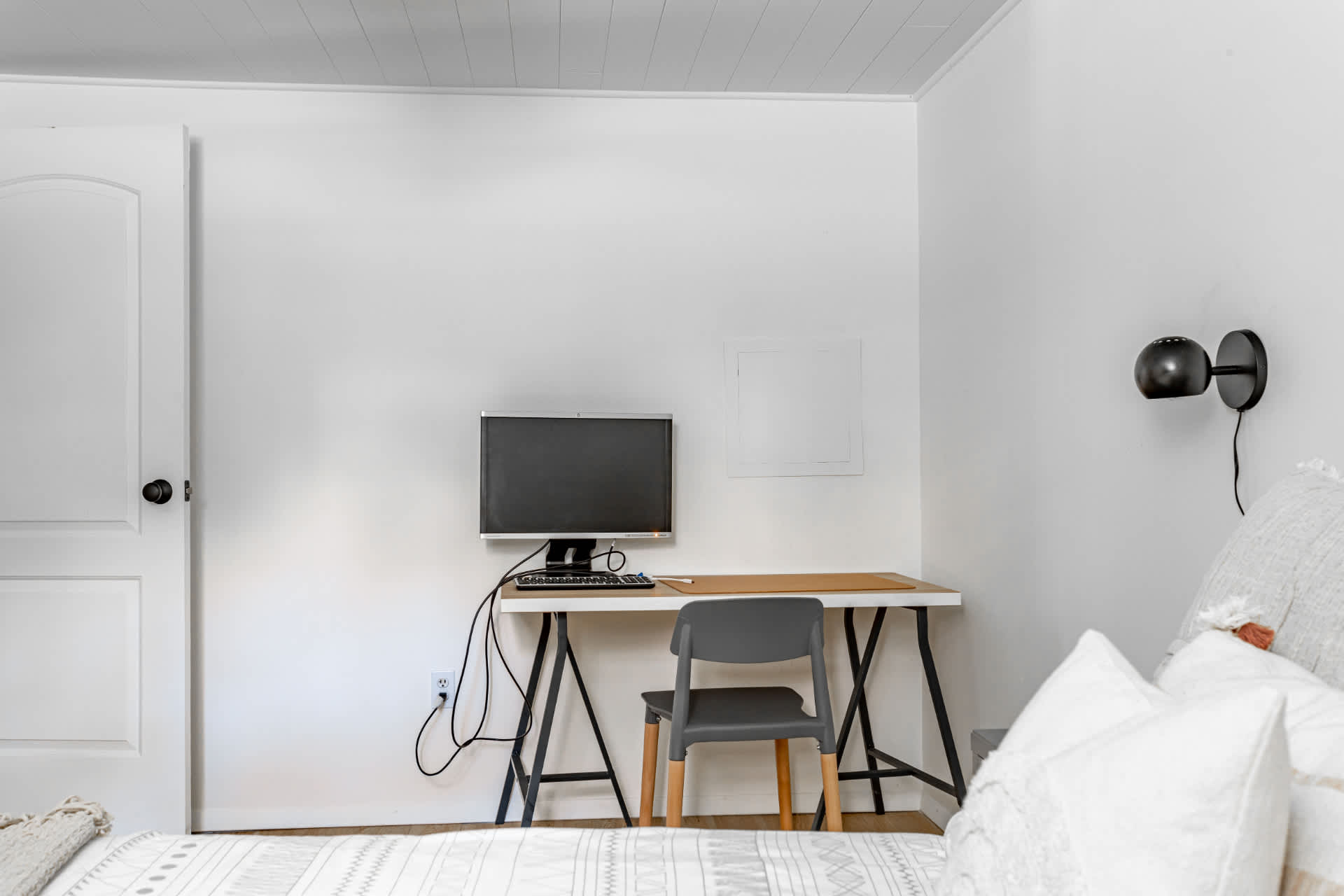 A dedicated workspace within a bedroom, featuring a black and white desk with a computer monitor and keyboard. A grey chair and a black wall sconce are also present.