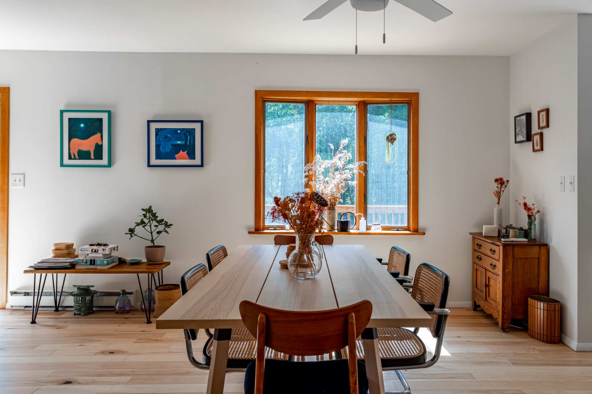 Bright dining room with a long light wood dining table surrounded by six chairs, a vase of dried flowers as a centerpiece, and a large window overlooking green trees, with light wood floors and white walls.