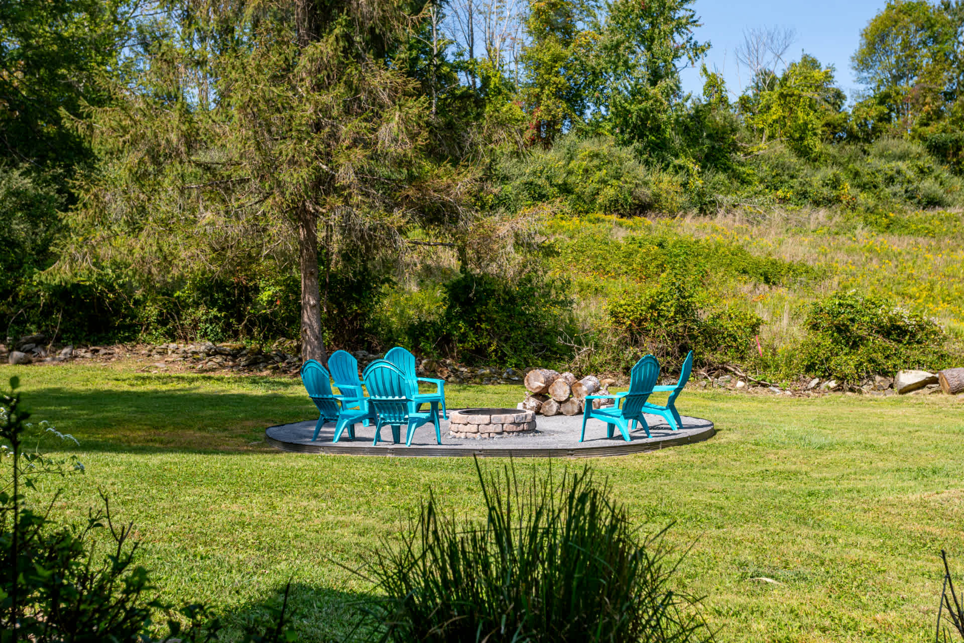 A circular fire pit area surrounded by several bright blue Adirondack chairs on a gravel base, set within a large grassy yard with mature trees and natural landscaping.