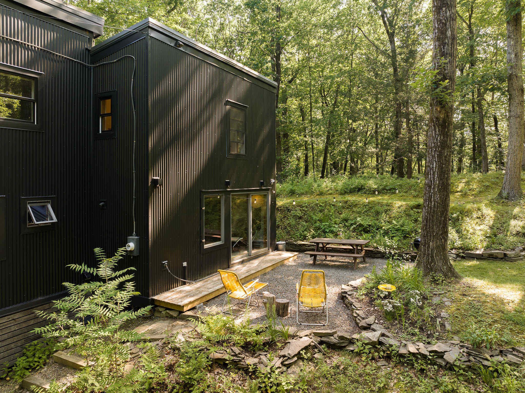 Modern black corrugated metal house exterior with a small wooden deck and two yellow outdoor chairs. A gravel patio area with a picnic table is visible, surrounded by lush green trees and natural landscaping.