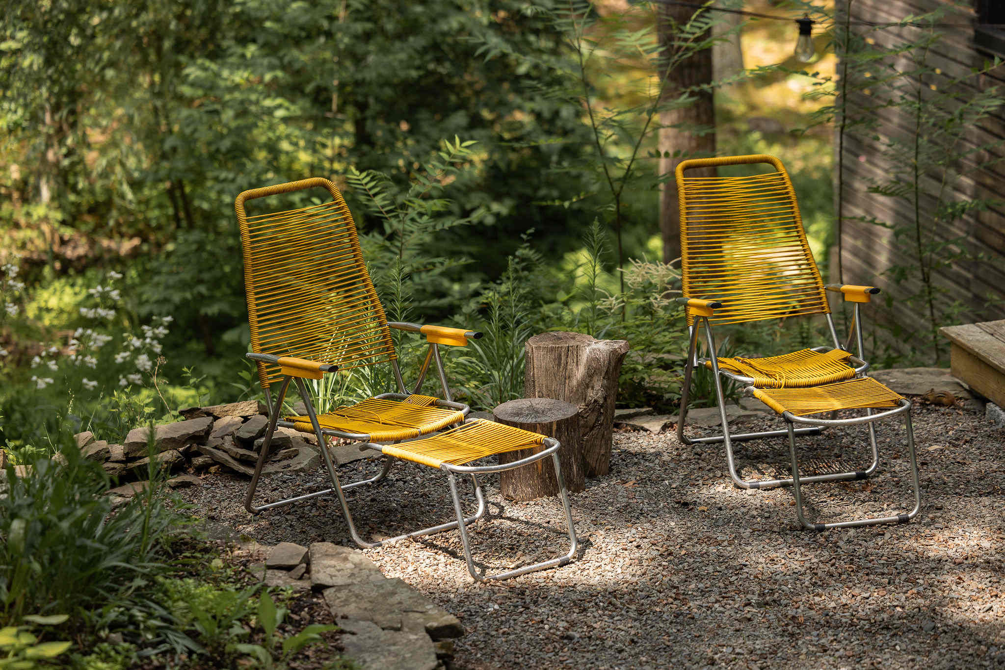 Relaxing outdoor seating area featuring two bright yellow slatted lounge chairs with footrests, placed on a gravel patch surrounded by lush greenery and trees.