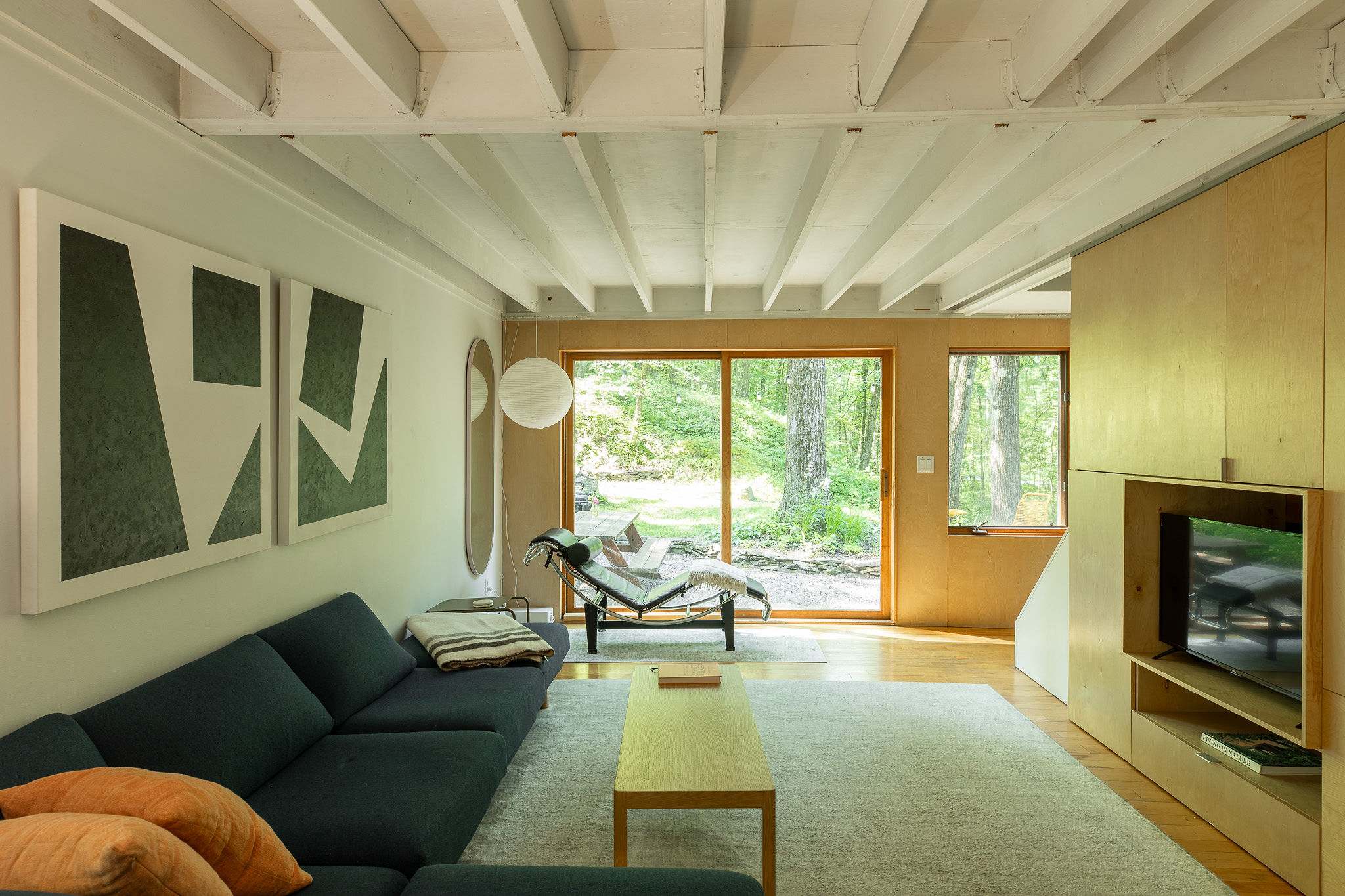 Expansive living room featuring exposed white ceiling beams, hardwood floors, and a large sliding glass door offering views of the surrounding forest. A dark blue sectional and a chaise lounge provide ample seating.