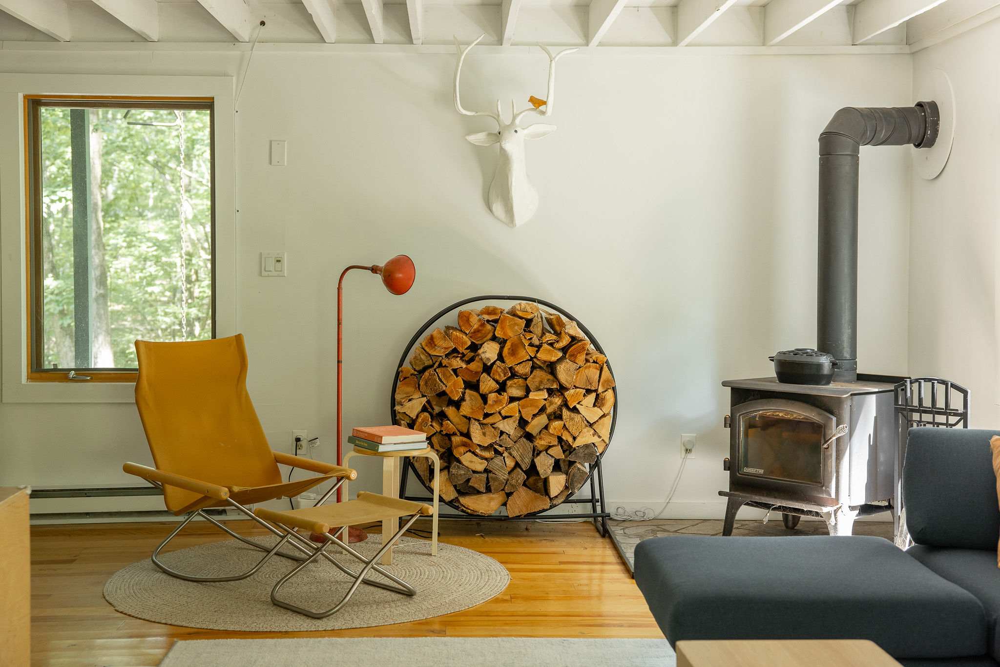 Detailed view of the living room's wood-burning stove area, featuring a black stovepipe and a circular metal rack filled with firewood. A white deer head mount adorns the wall above a yellow armchair.