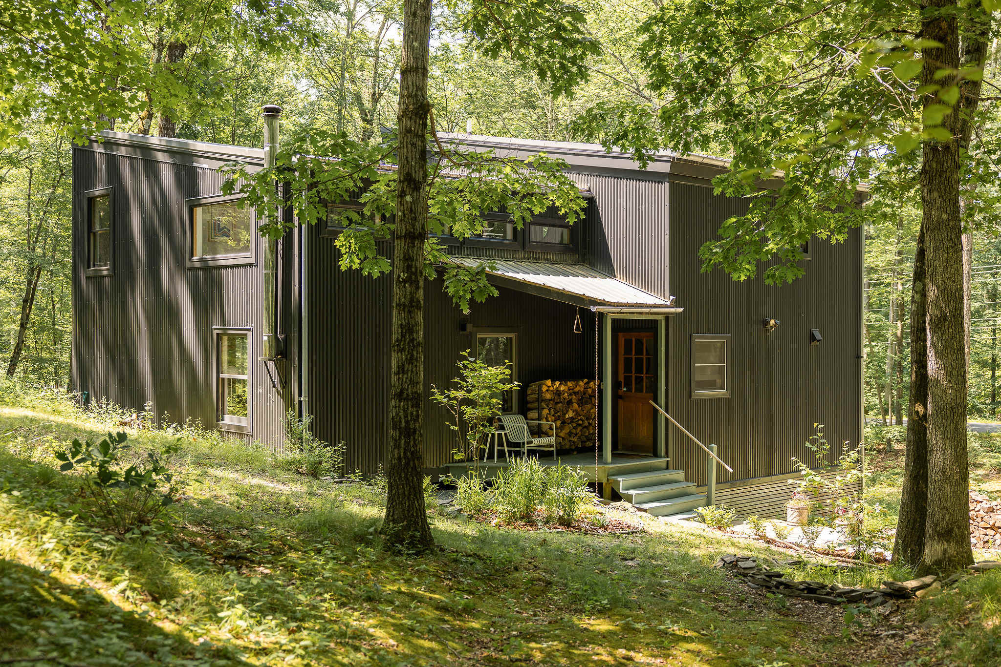 Contemporary black corrugated metal house with a covered front porch, wooden steps, and a stack of firewood, surrounded by dense green trees.