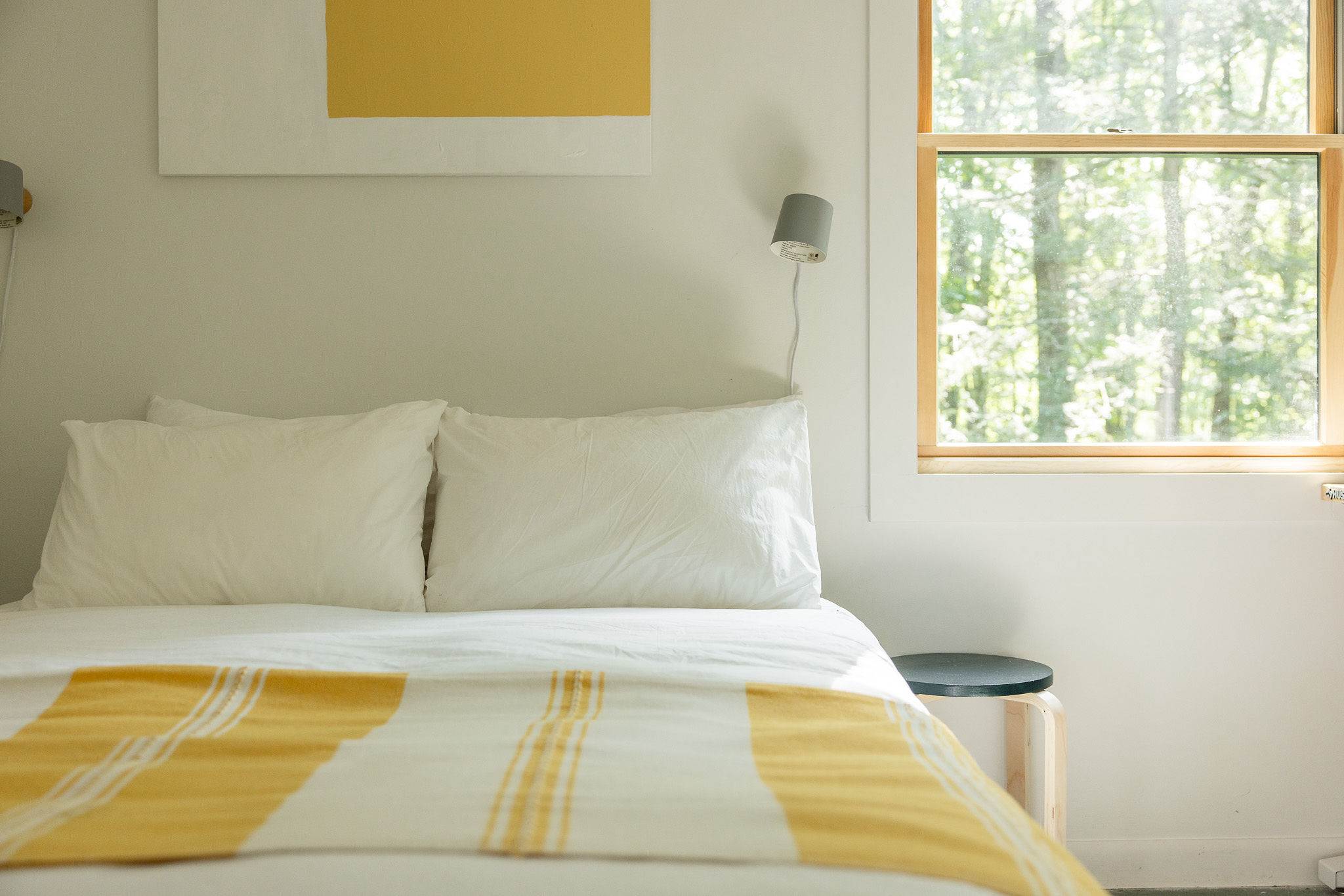Close-up of a white bed with two pillows and a yellow and white striped throw blanket. A small round side table with a dark top is next to the bed, and a grey wall-mounted lamp is visible above.