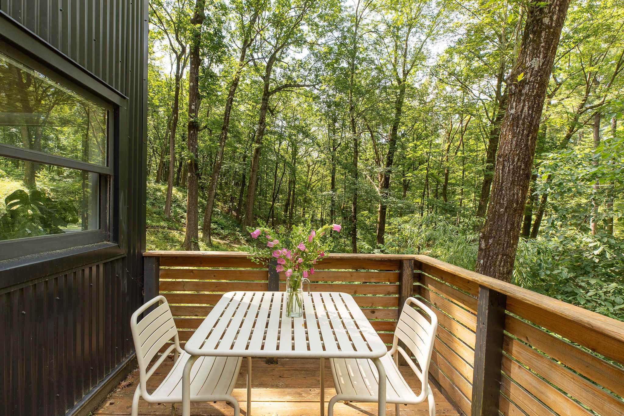 Outdoor deck with a white slatted table and two matching chairs, enclosed by a wooden railing, offering a serene dining spot amidst the dense green forest.