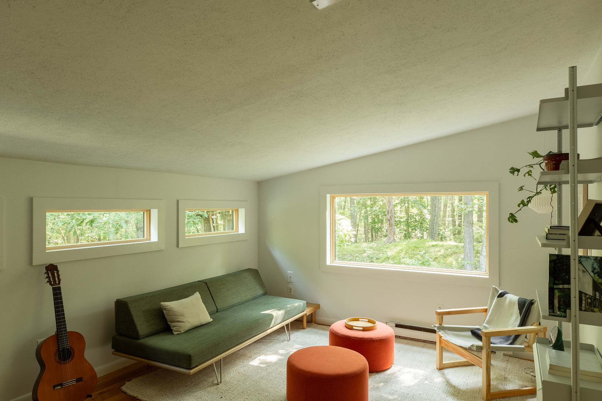 Bright secondary living space with a green futon-style sofa, two orange ottomans, a light wood armchair, and multiple windows providing views of the surrounding trees.