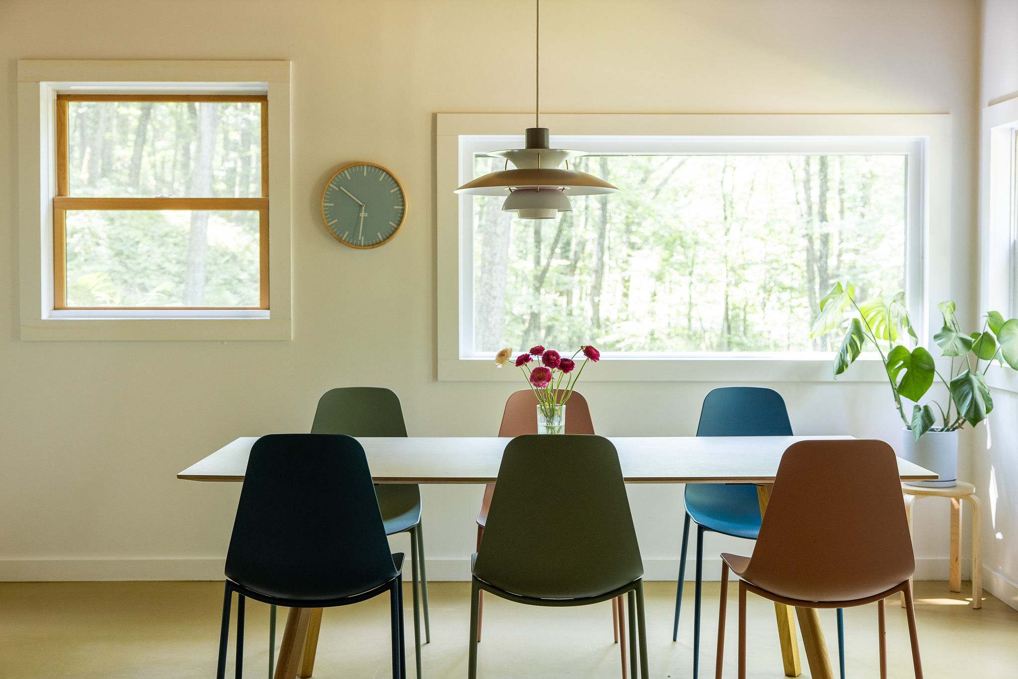 Close-up view of the dining area, showcasing a white rectangular table adorned with flowers and surrounded by a variety of colorful, modern dining chairs. Large windows offer a lush green outdoor view.