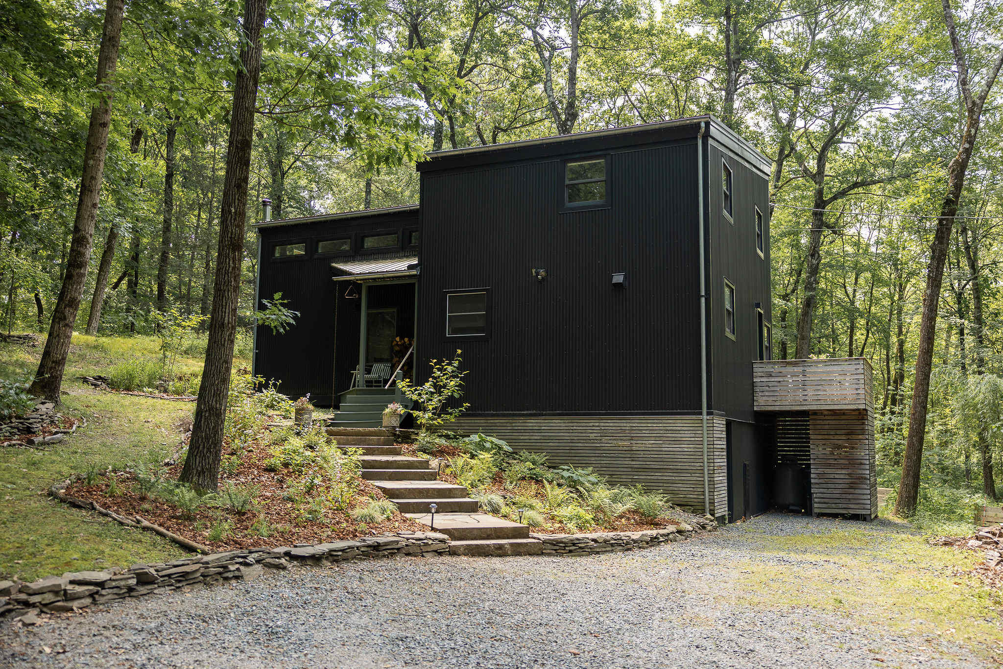 Modern black corrugated metal house with a gravel driveway leading to stone steps and a covered entrance, surrounded by a verdant forest.