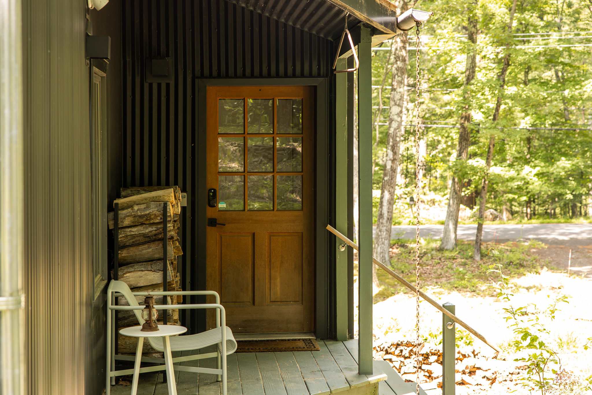 Inviting front porch with a wooden paneled door, a stack of firewood, and a white outdoor chair, set against the dark corrugated siding of the house.