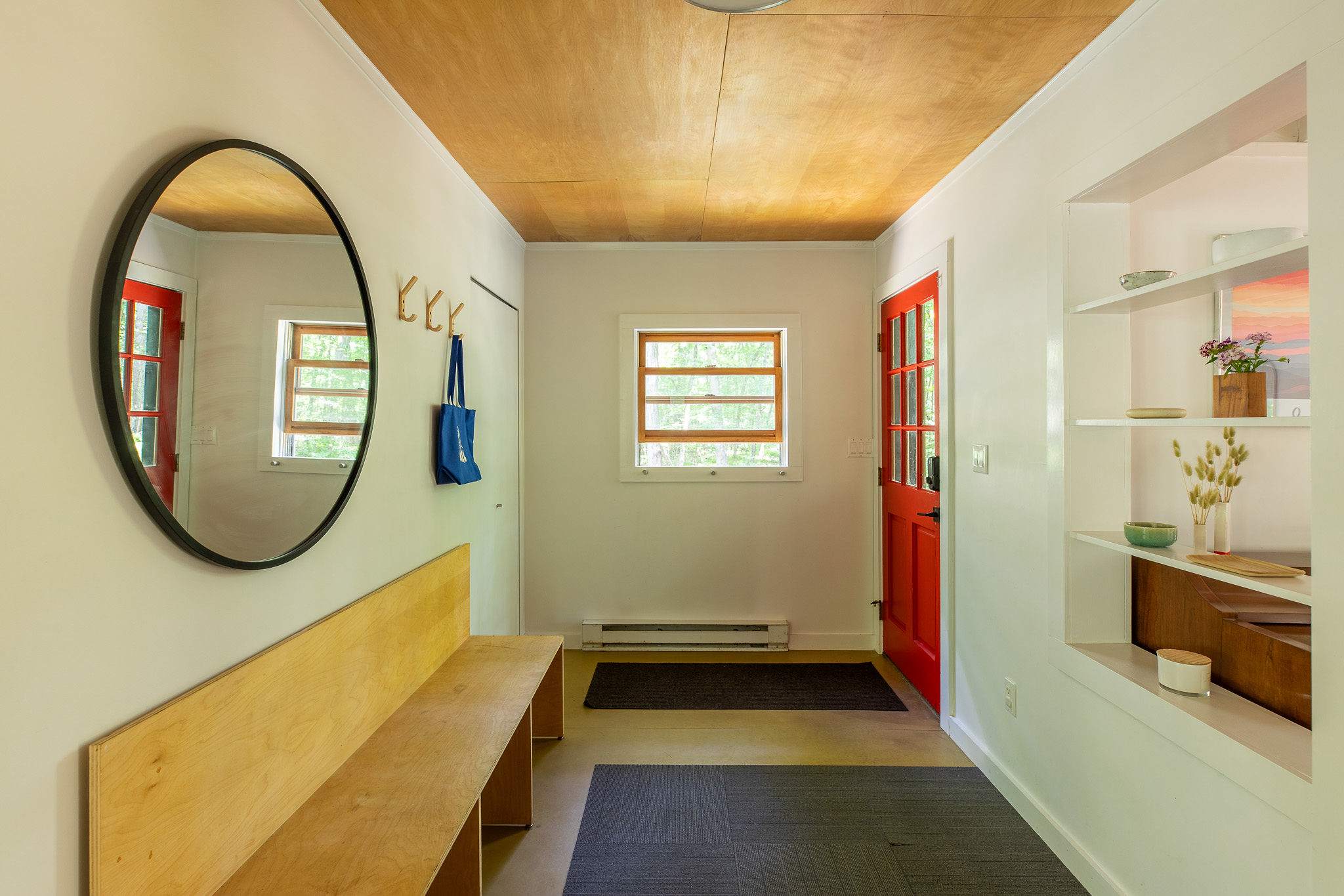 Bright and minimalist entryway featuring a long wooden bench, a round black mirror, and a vibrant red front door, with a light wood ceiling and white walls.