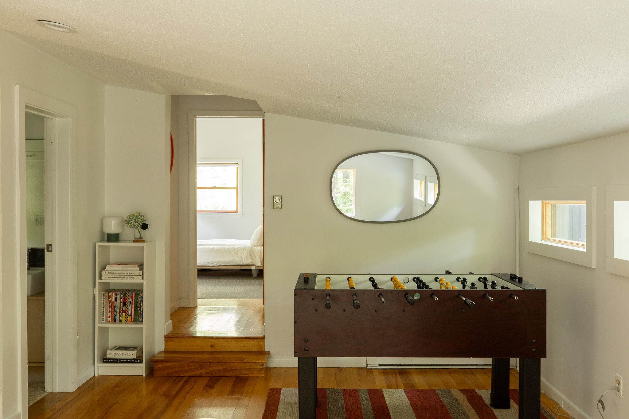 Bright game room or landing area featuring a foosball table on a striped rug, with a decorative oval mirror on the wall. A small bookshelf holds various items, and a doorway leads to another room.