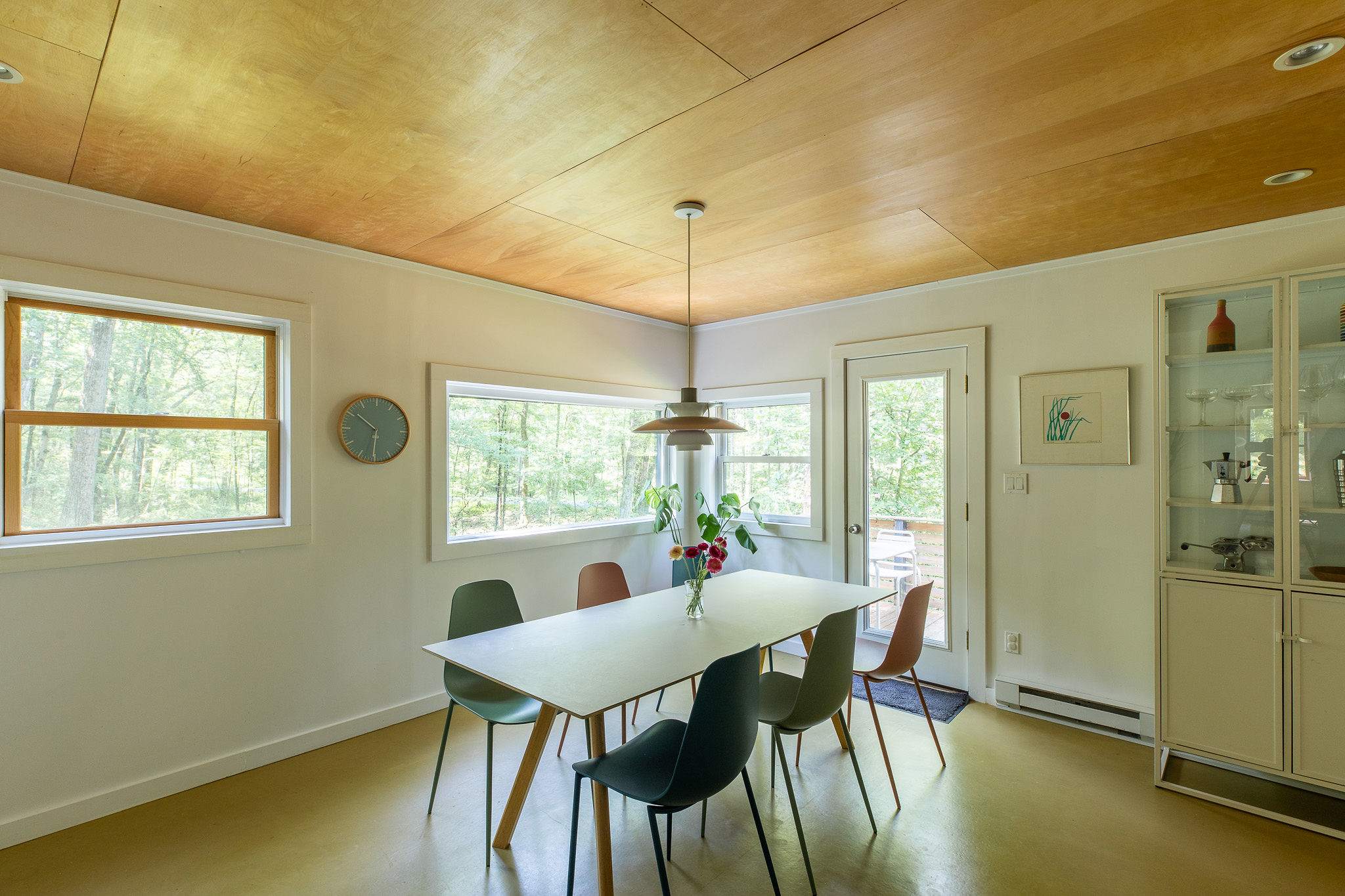 Spacious dining area featuring a long white table with a mix of modern chairs in muted tones, illuminated by natural light from large windows. A wood-paneled ceiling adds warmth to the space.