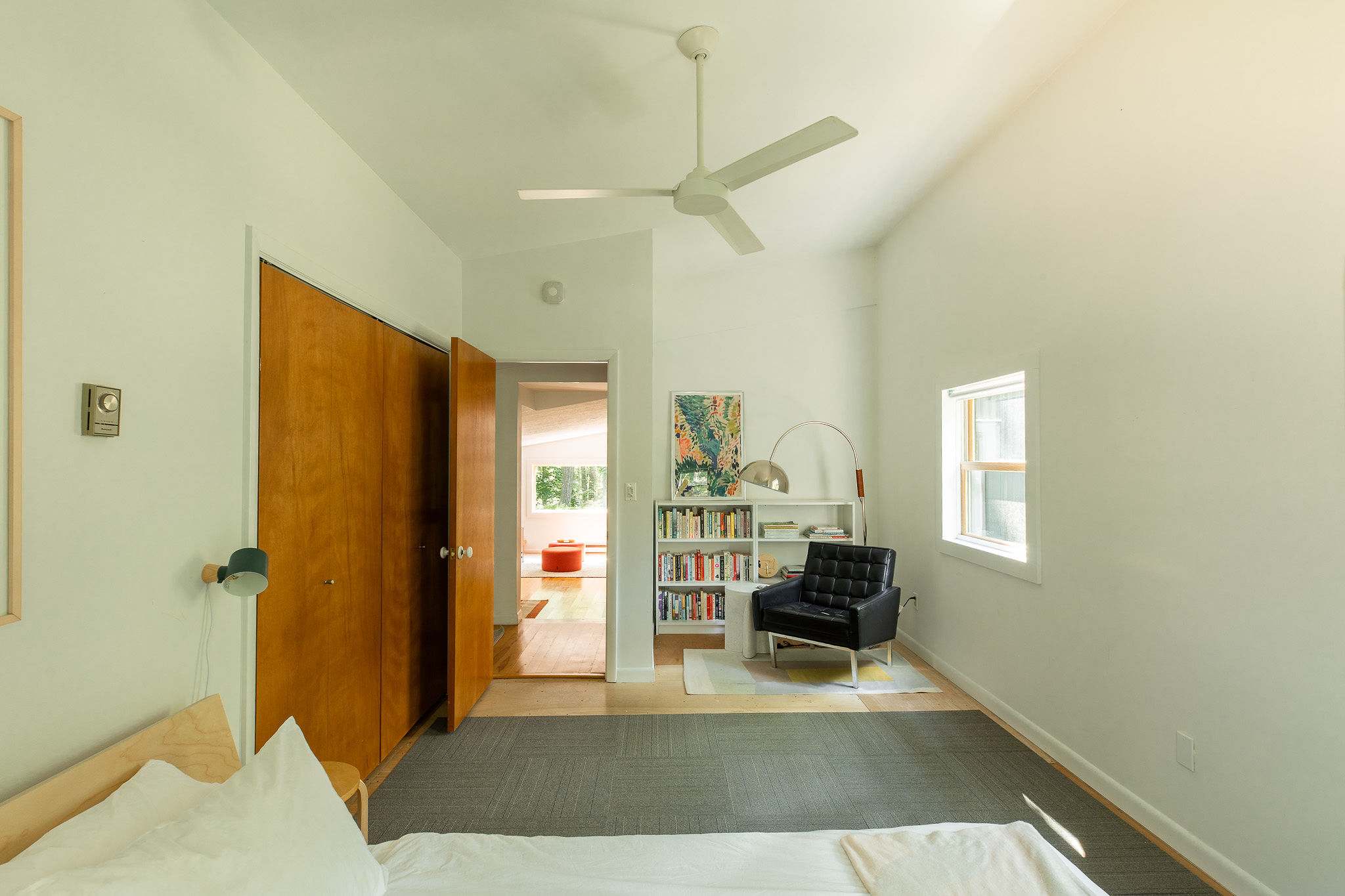 Spacious bedroom with a white bed, a sitting area featuring a black armchair and a bookshelf, and a ceiling fan. A doorway leads to another room, and a large closet with wood doors is visible.