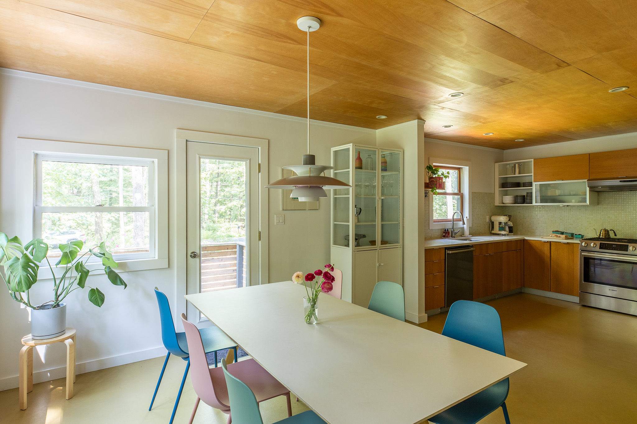 Bright dining area with a long white table surrounded by colorful modern chairs, set under a warm wood-paneled ceiling. The kitchen is visible in the background, maintaining a cohesive design.