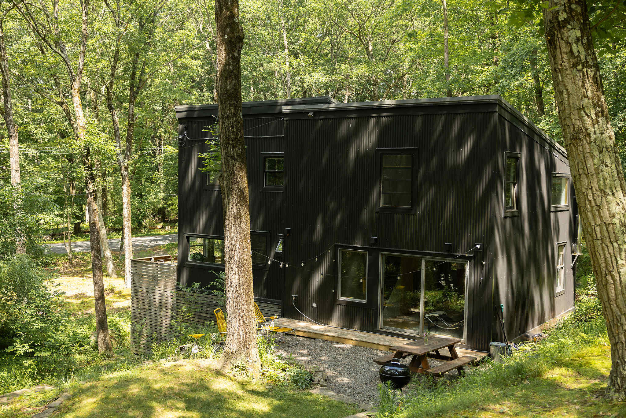 Modern black corrugated metal house nestled in a lush green forest, featuring a small wooden deck with a picnic table, BBQ grill, and string lights overhead.