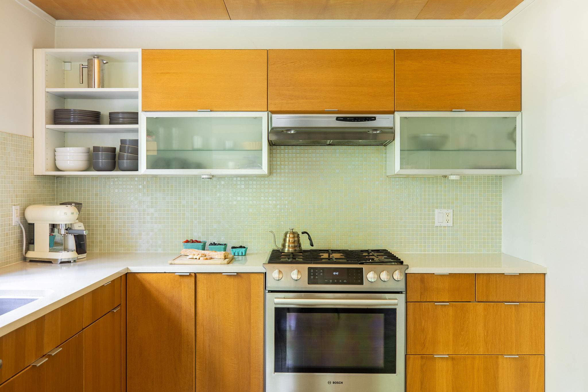 Modern kitchen featuring light wood cabinetry, white countertops, and a light green mosaic tile backsplash. Stainless steel appliances include a gas range and range hood, with open shelving displaying dishes.