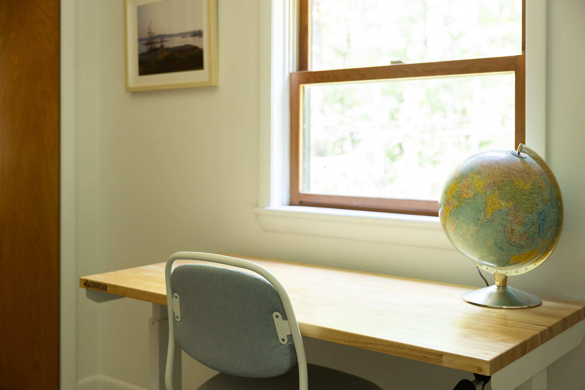 Close-up of a light wood desk with a grey office chair and a vintage-style globe, positioned in front of a window. The window offers a bright view of lush green trees outside.