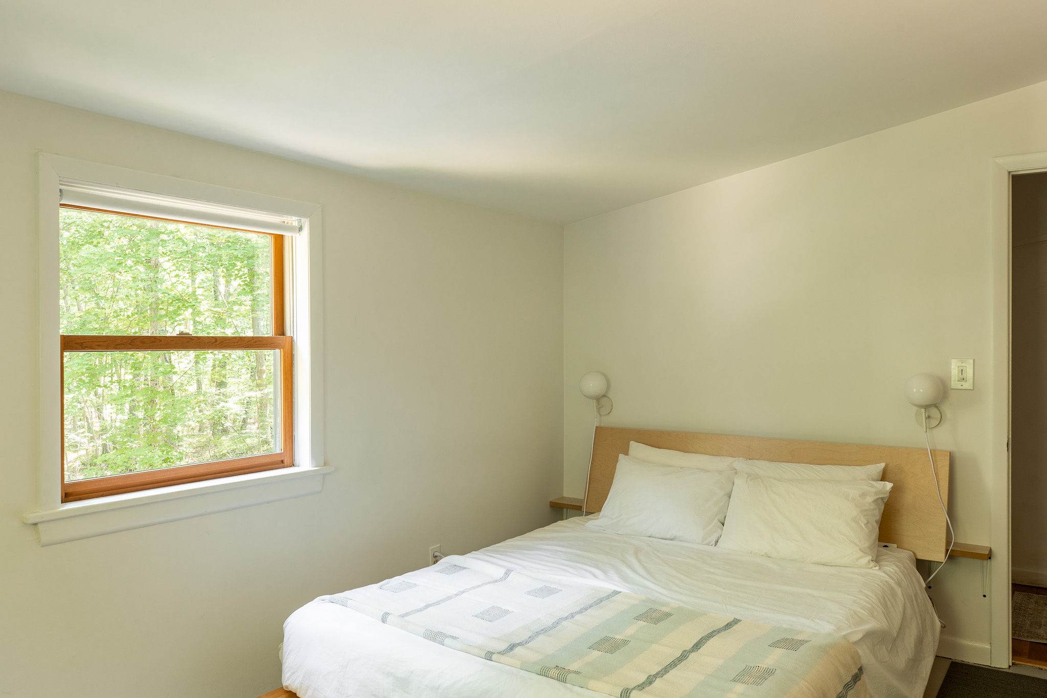 Close-up of a white bed with a light wood headboard and a light blue and white patterned throw blanket. A window to the left provides natural light and a view of green trees.