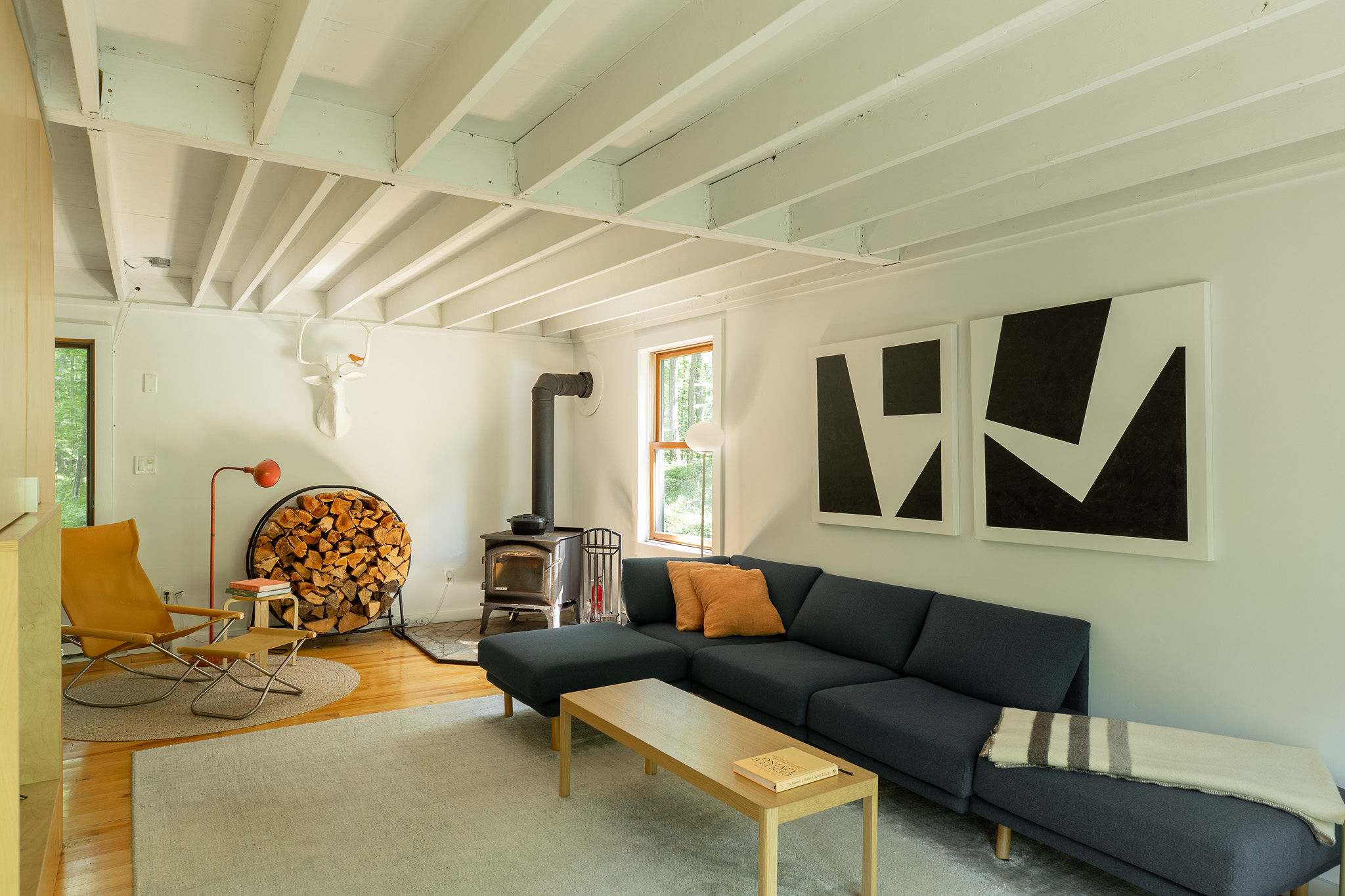 Cozy living room with exposed white ceiling beams, hardwood floors, and a dark blue sectional sofa. A wood-burning stove sits beside a circular firewood rack, complemented by a yellow armchair and modern artwork.