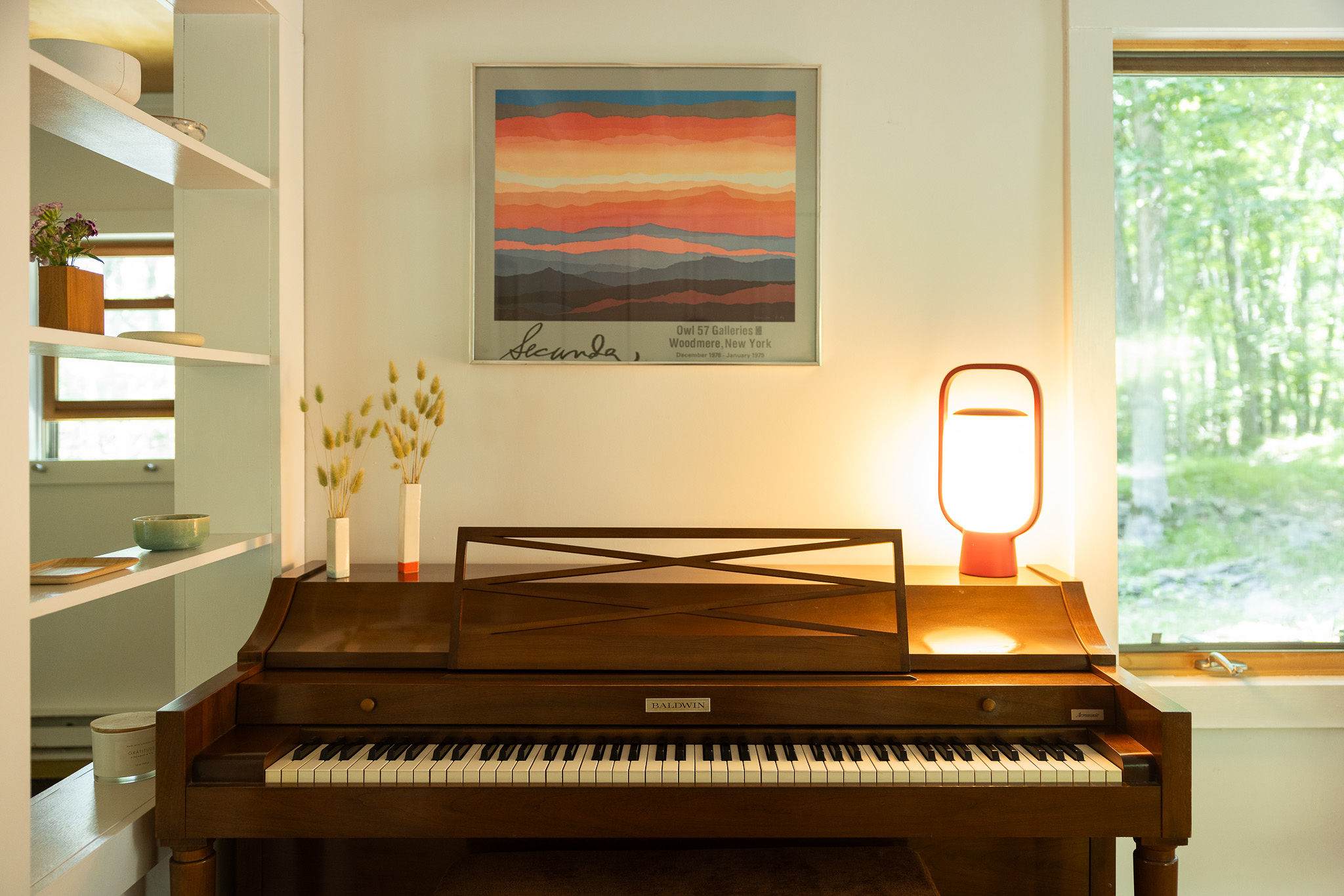 A classic wooden piano is positioned against a white wall, beneath a framed abstract artwork. A modern, minimalist table lamp provides soft illumination to the space.