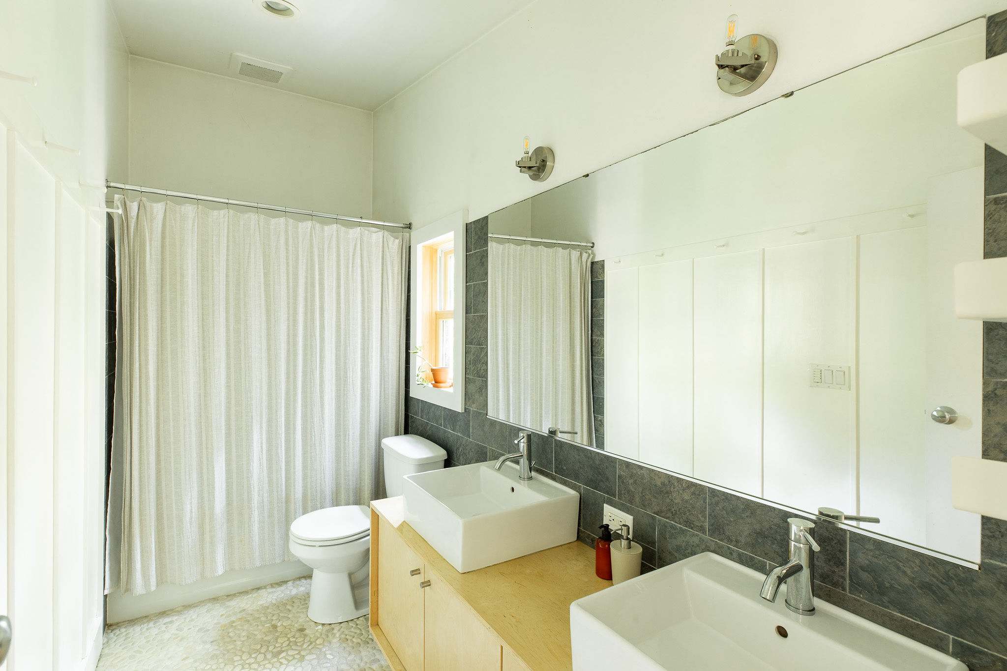Modern bathroom featuring a double vanity with square white sinks set on a light wood cabinet, against a dark grey tiled backsplash. A large mirror spans the wall, and a shower with a white patterned curtain is visible.
