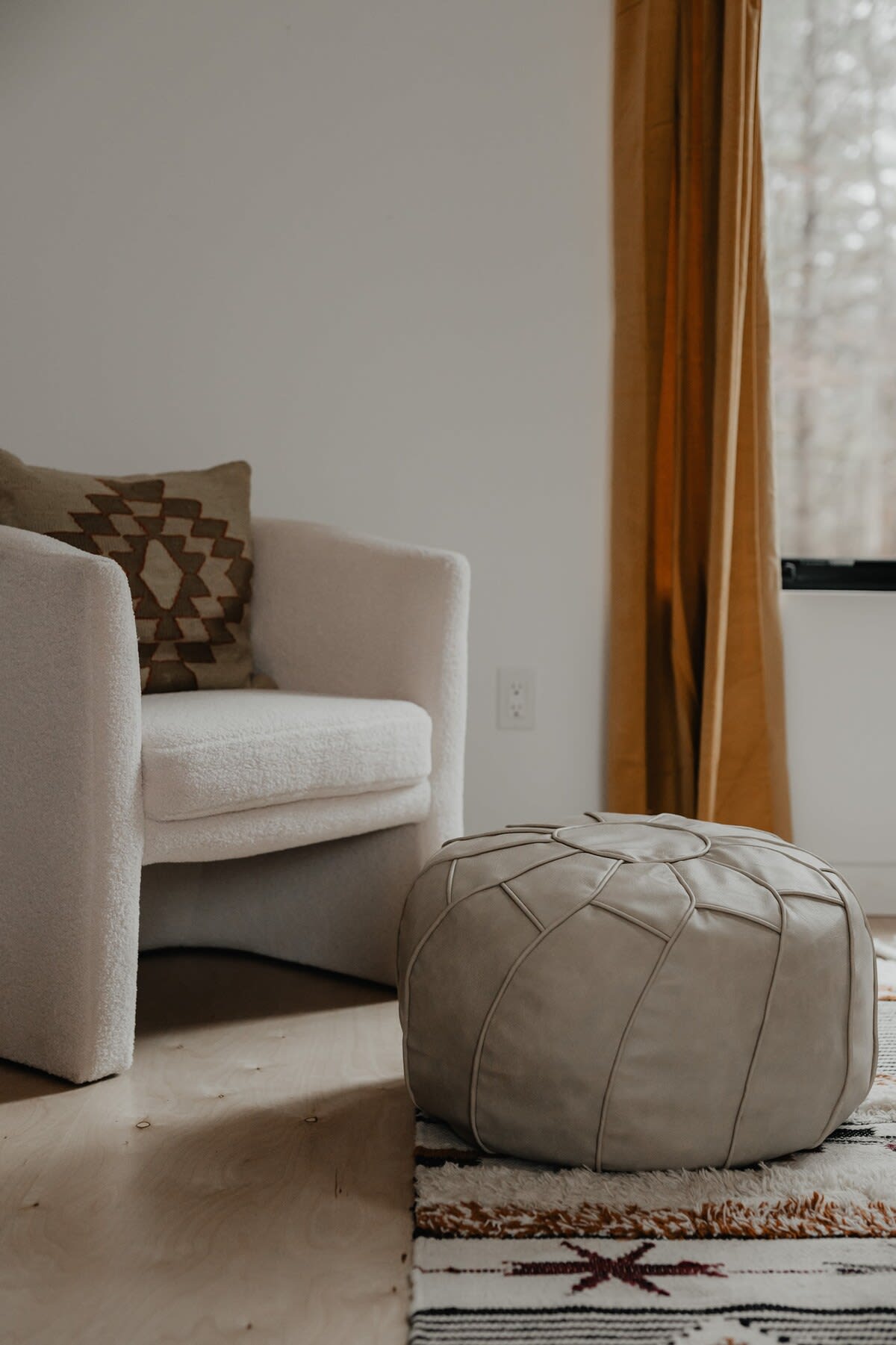 A cozy corner in a bedroom featuring a textured cream armchair with a patterned throw pillow, and a light-colored leather pouf, set on a patterned rug next to a window with mustard yellow curtains.